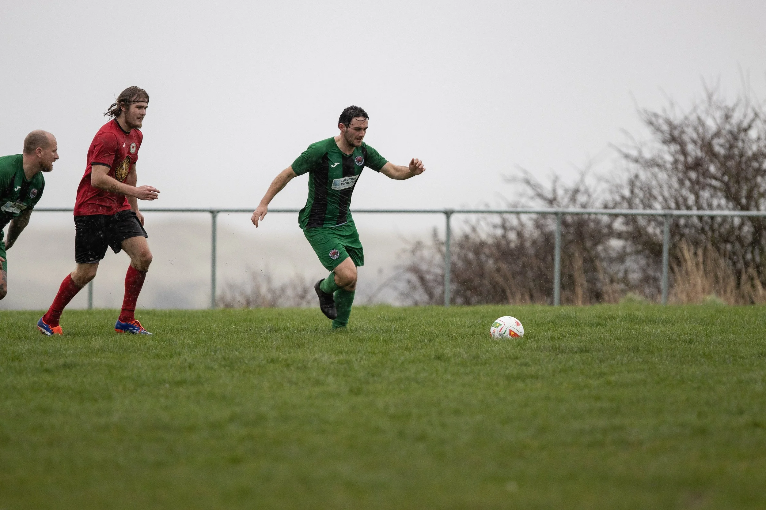 Soccer players running on a grassy field during a match, with one player in a green and black uniform approaching the ball while the others in red and green chase behind.