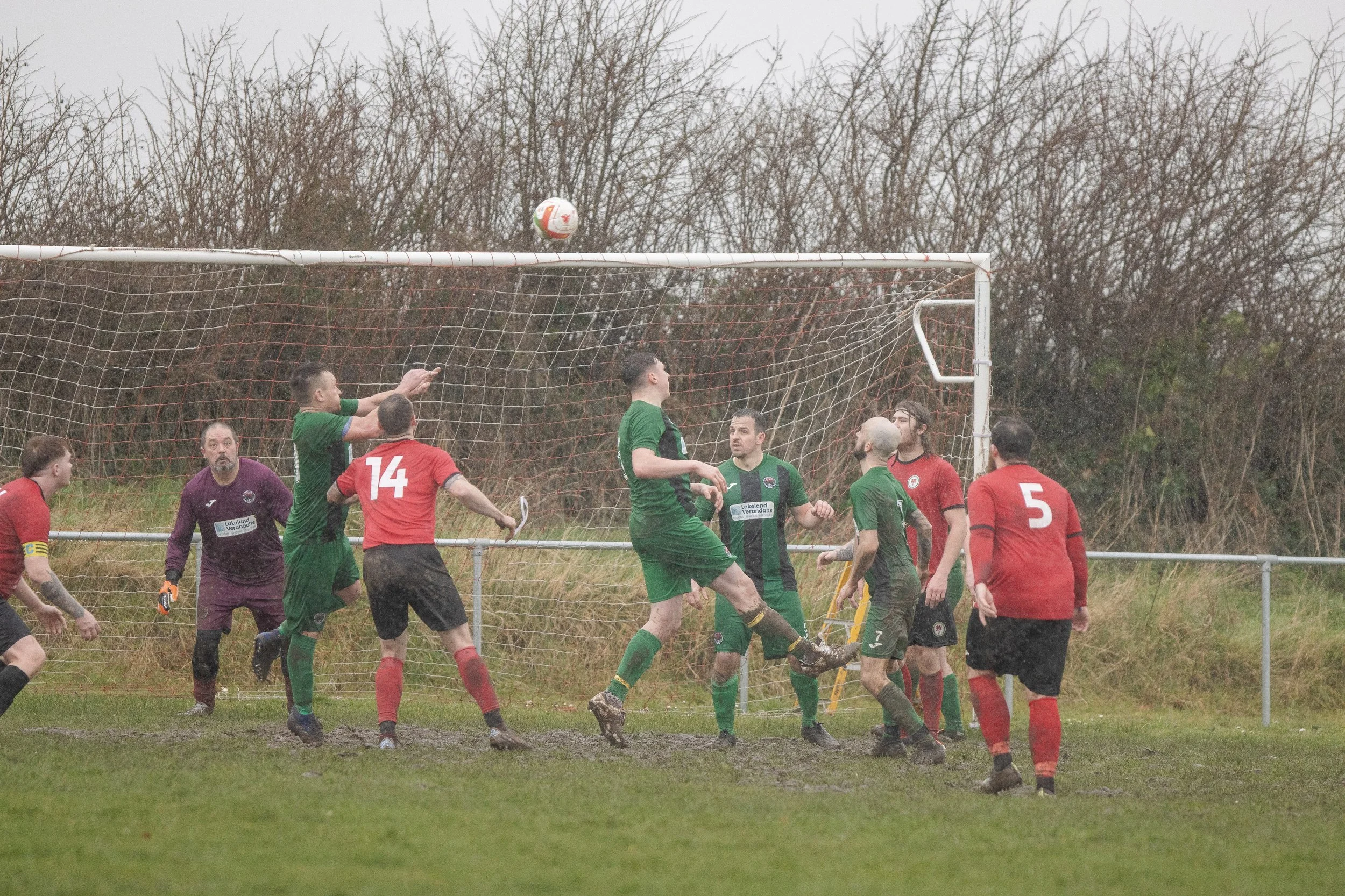 A soccer game on a muddy field with players from both teams competing in front of the goal. The players are wearing red and green jerseys, with some jumping and others watching the ball in the air. The background shows leafless trees and an overcast 