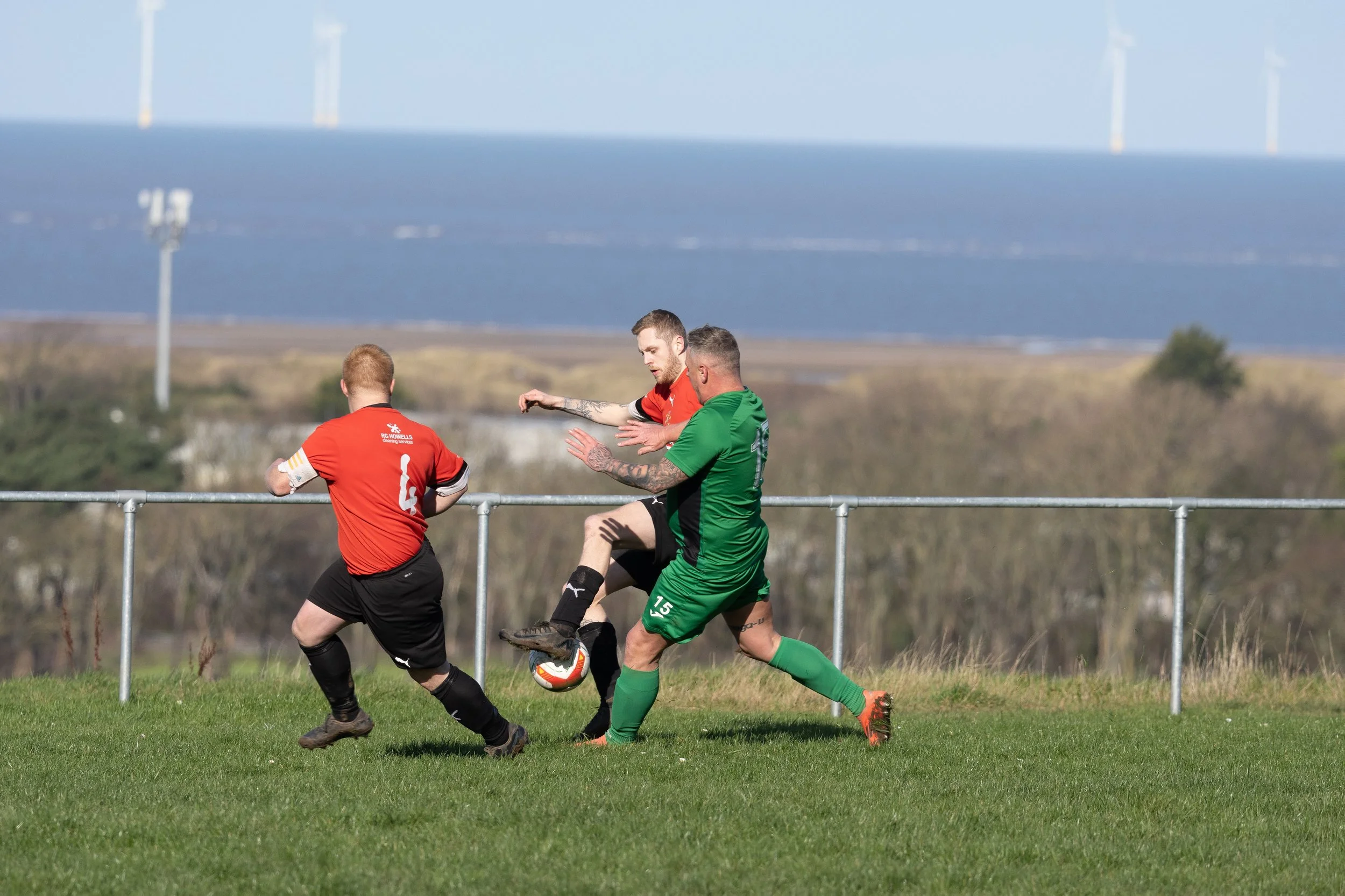 Three soccer players in action on a grass field with a scenic background of open land, trees, and wind turbines in the distance. Two players are wearing red jerseys, and one is wearing green.