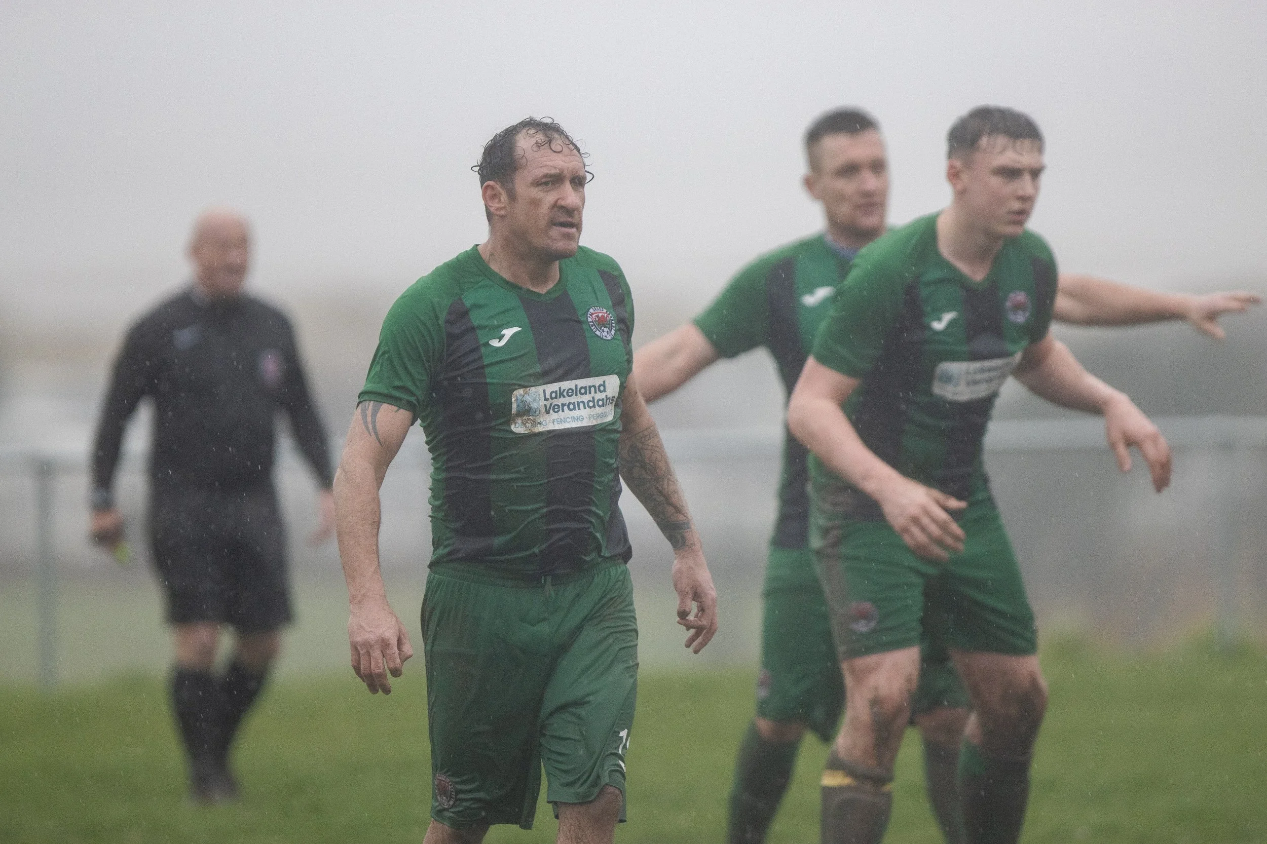 Soccer players in green jerseys standing on a field in rainy weather, with one player looking serious and others with outstretched arms.