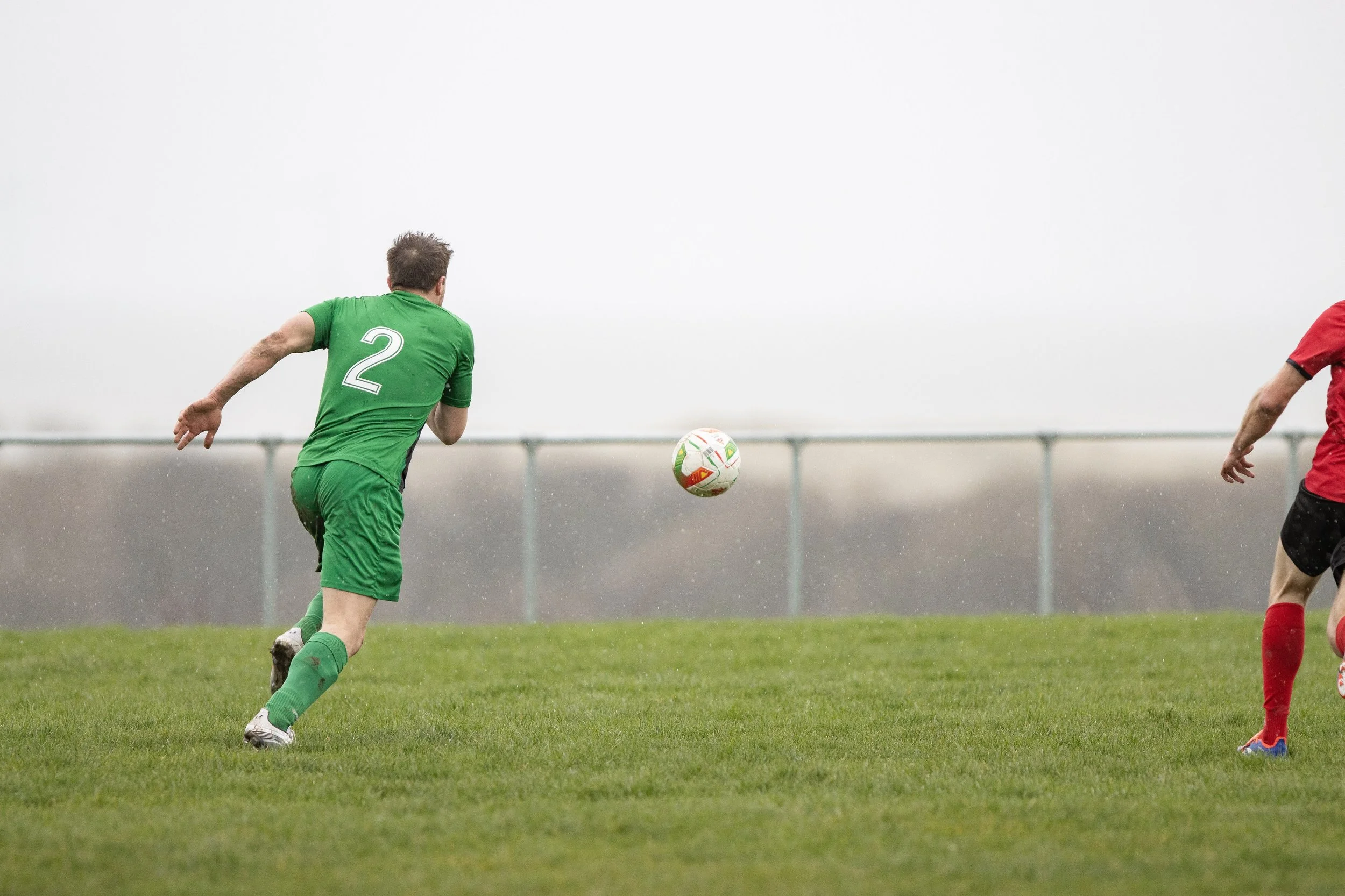 A soccer player in a green uniform with the number 2 on his back running towards a soccer ball on a grassy field, with another player partially visible on the right, under a cloudy sky.