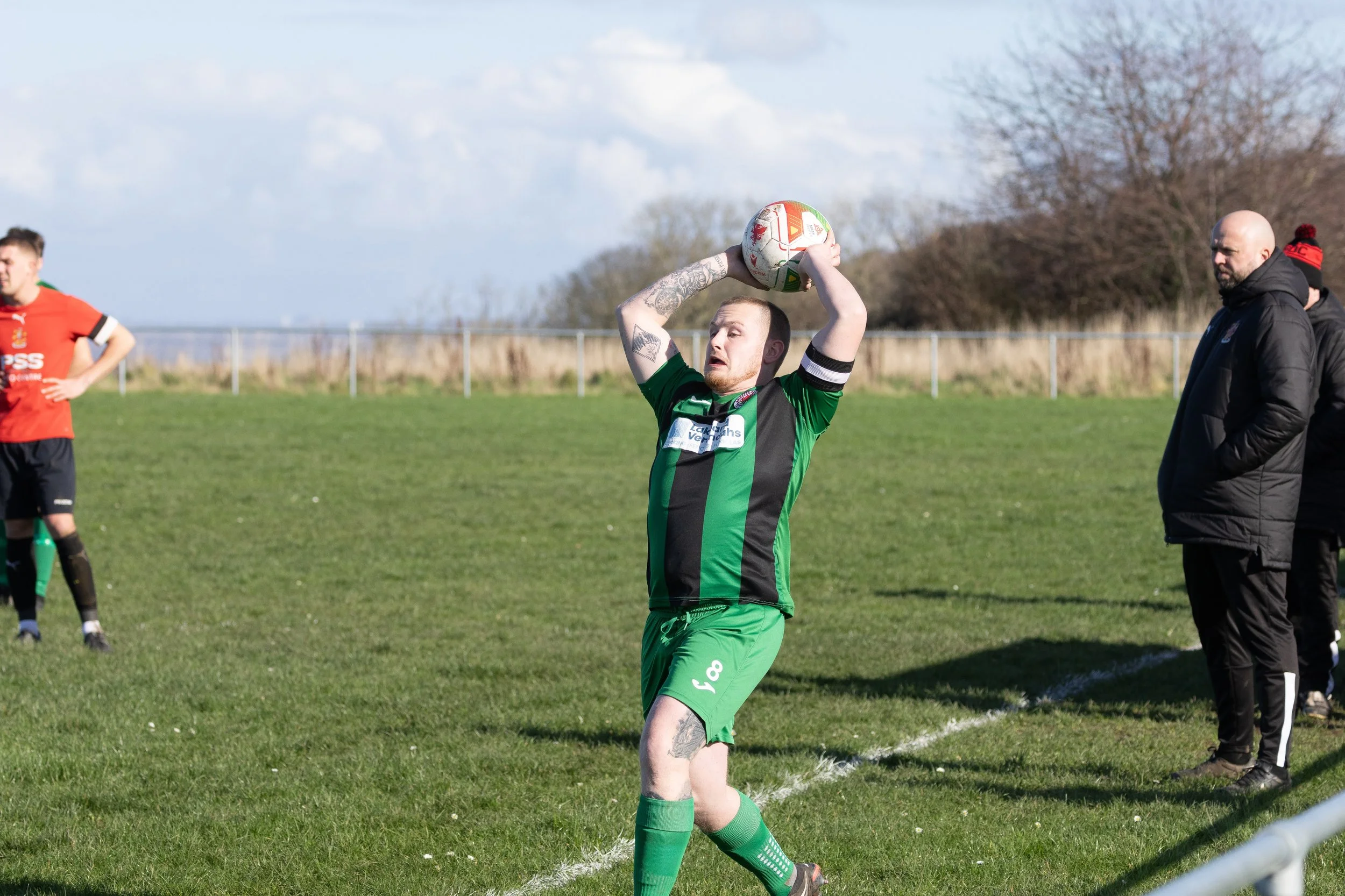 A soccer player in a green and black uniform prepares to throw a ball during a match on a grassy field, with two spectators or coaches standing on the sideline watching.