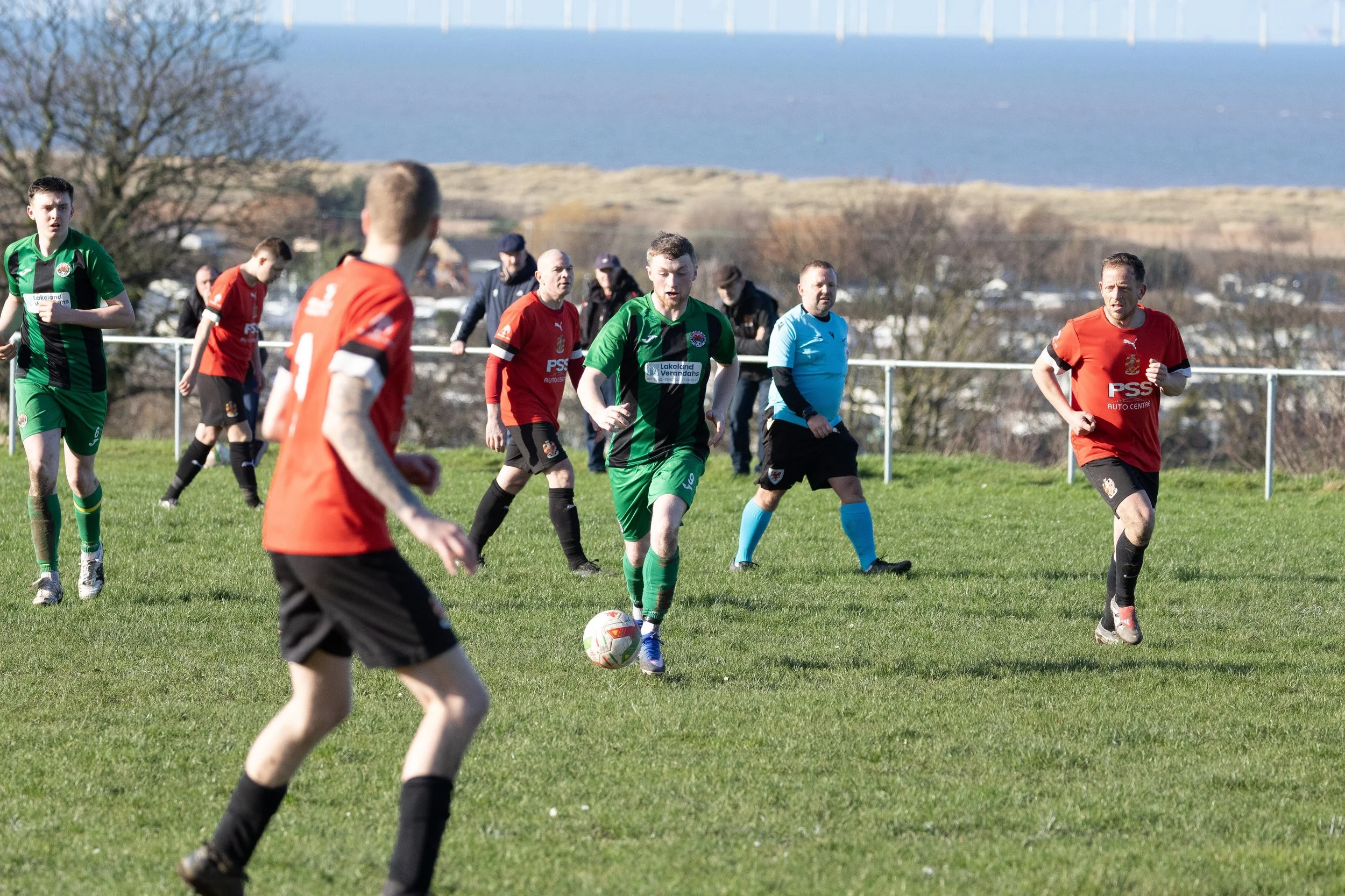 Soccer match with players in red and green jerseys on a grassy field, with a referee in blue overseeing the game. The background shows trees, a fence, and a distant body of water under a clear sky.
