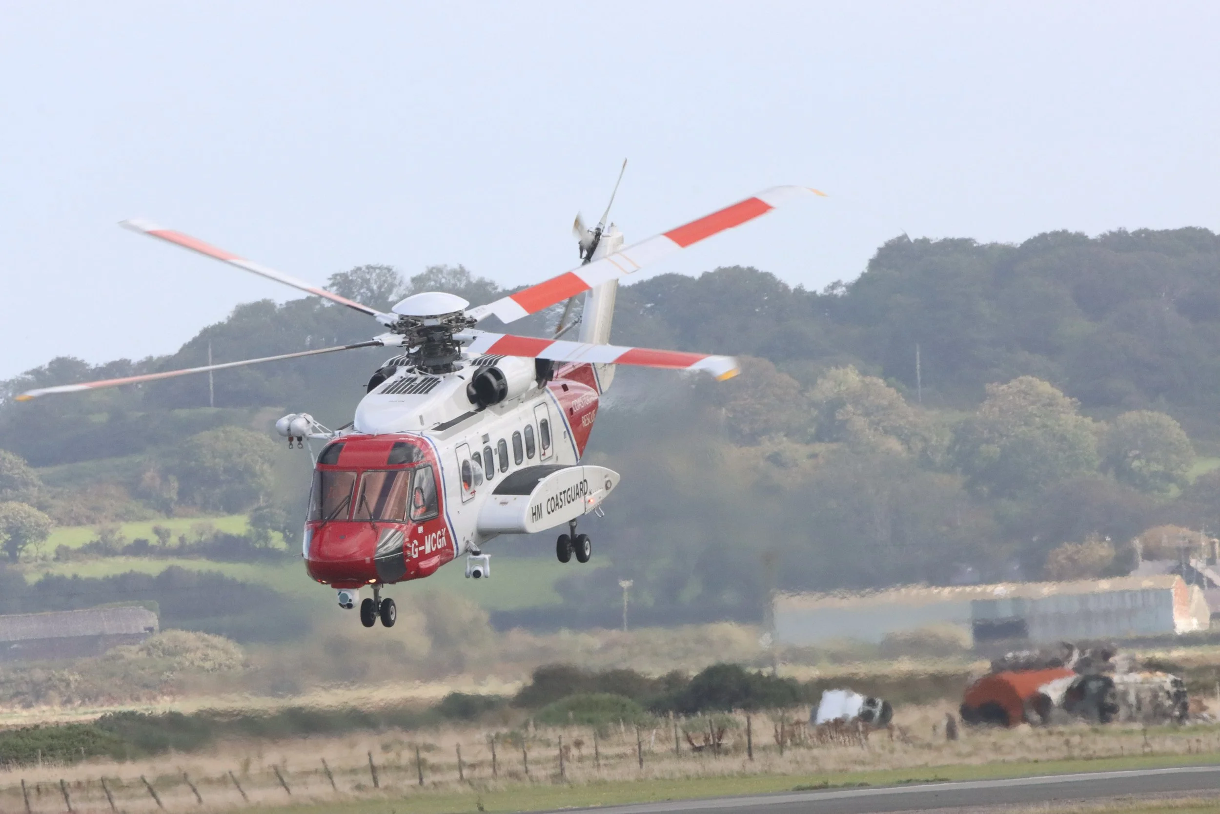 A white and red coast guard helicopter flying low over a field with fallen burning cars and a fence, with trees in the background.
