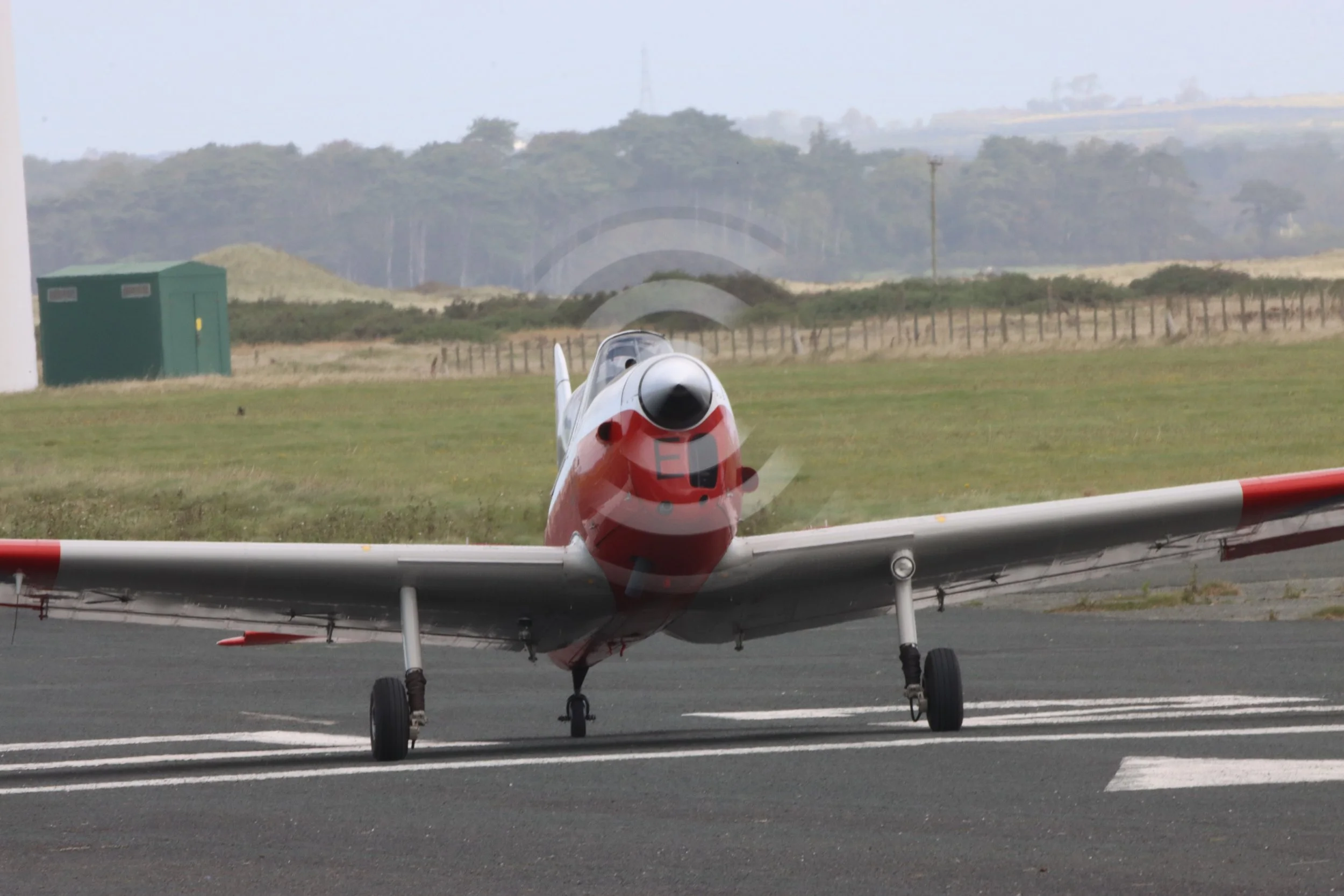 A vintage propeller airplane on a runway at an airfield, with grassy fields and trees in the background.
