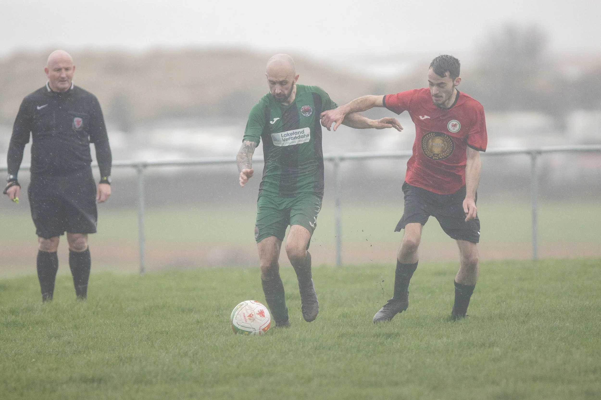 Two soccer players competing for the ball on a rainy field, with a referee observing in the background.