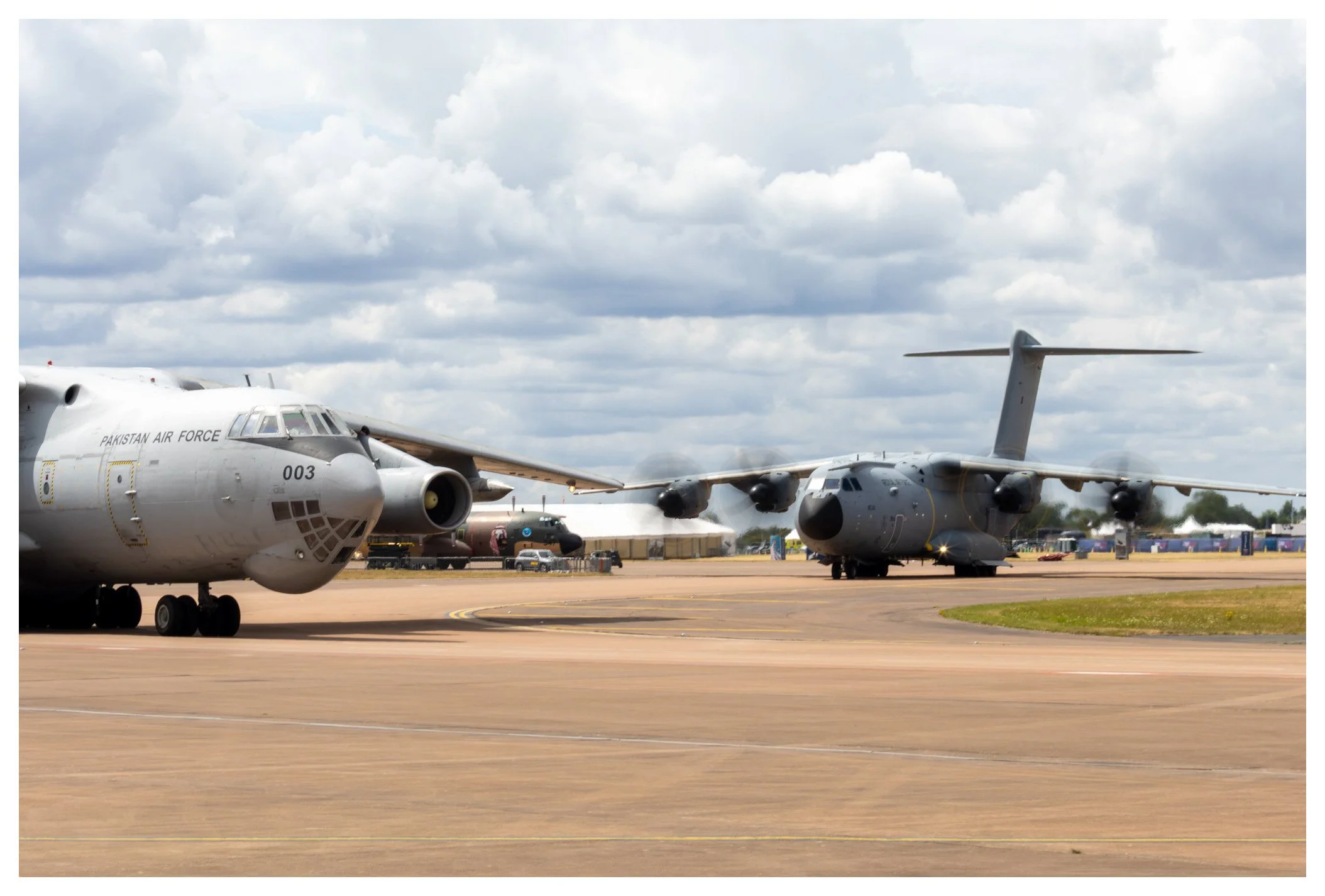 Two military cargo planes on the tarmac at an airbase, with a cloudy sky overhead.