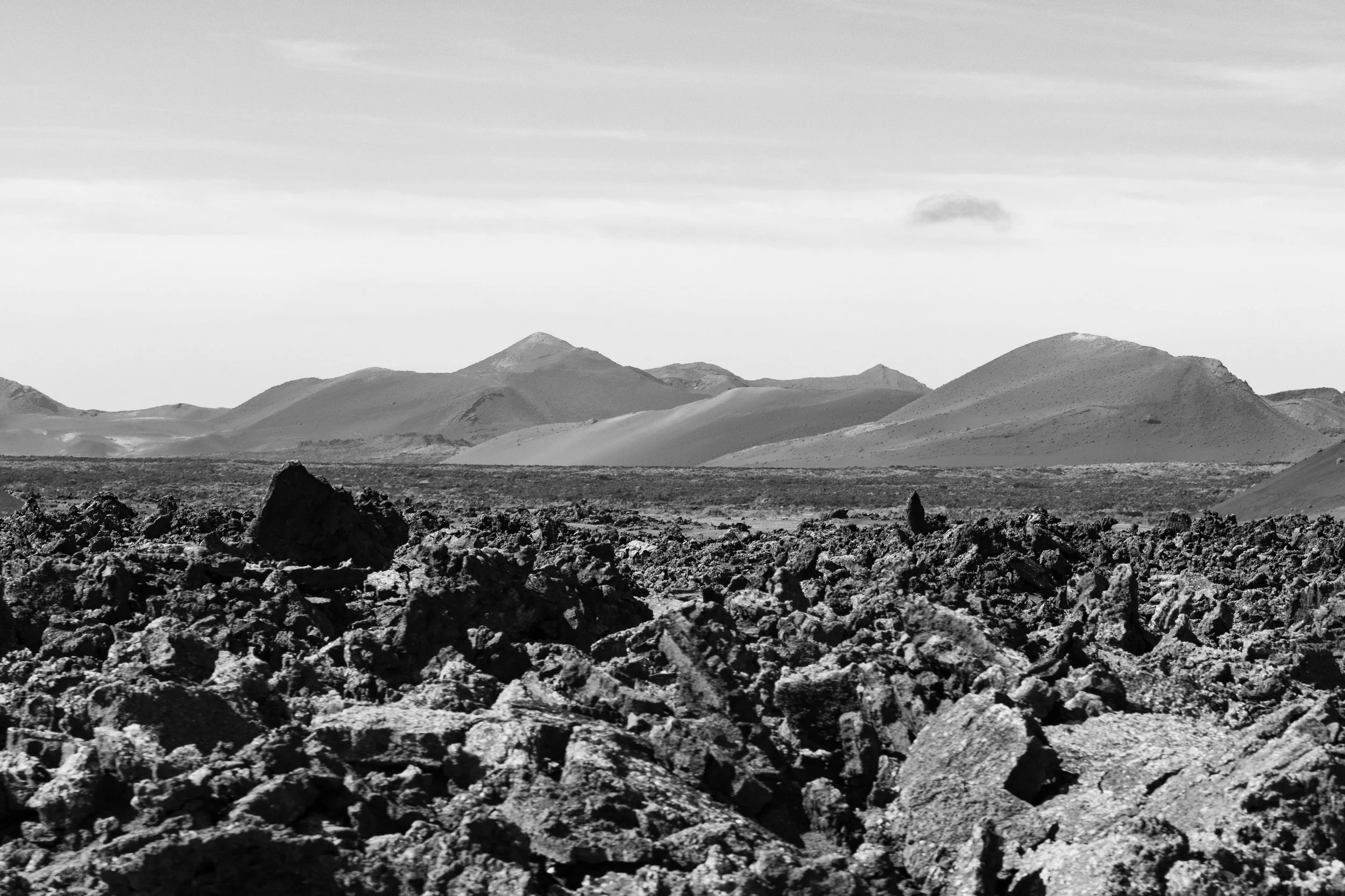 Black and white landscape of a volcanic terrain with rough lava rocks in the foreground and mountain ranges in the distance under a cloudy sky.