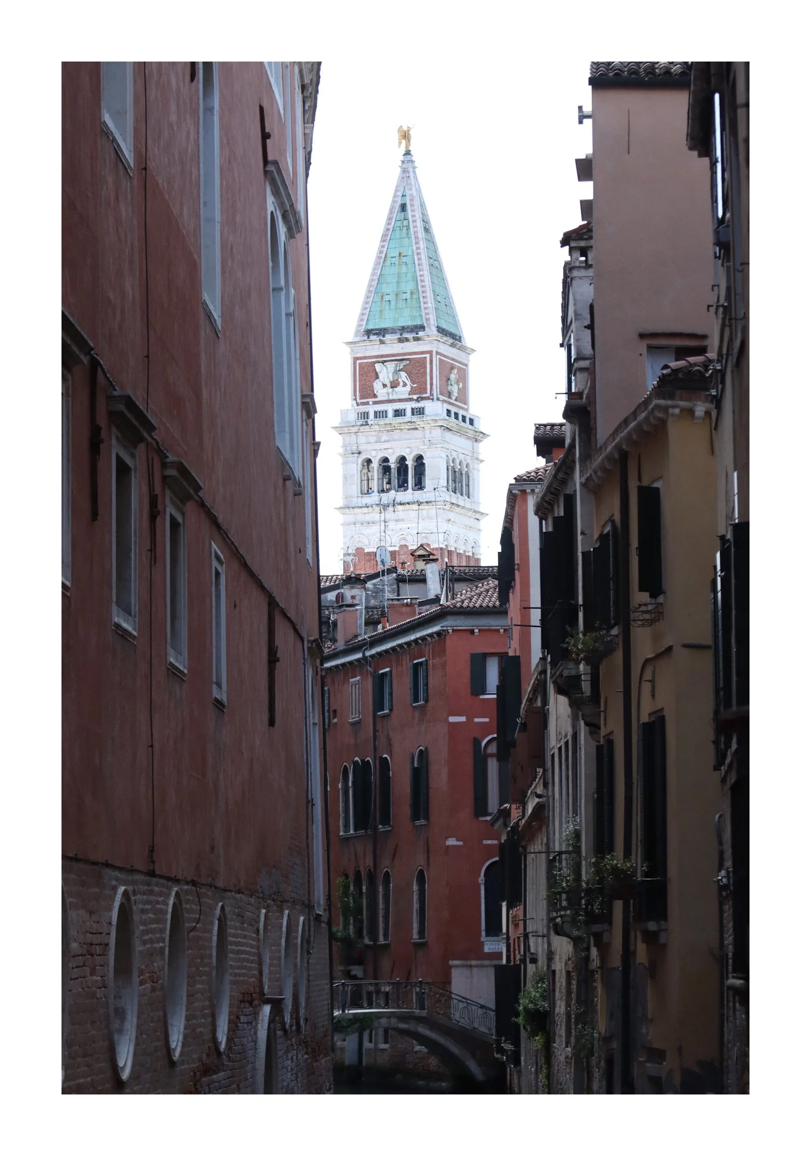 View of a narrow canal street with colorful old buildings on both sides and the bell tower of St. Mark's Campanile in Venice, Italy, visible in the background.