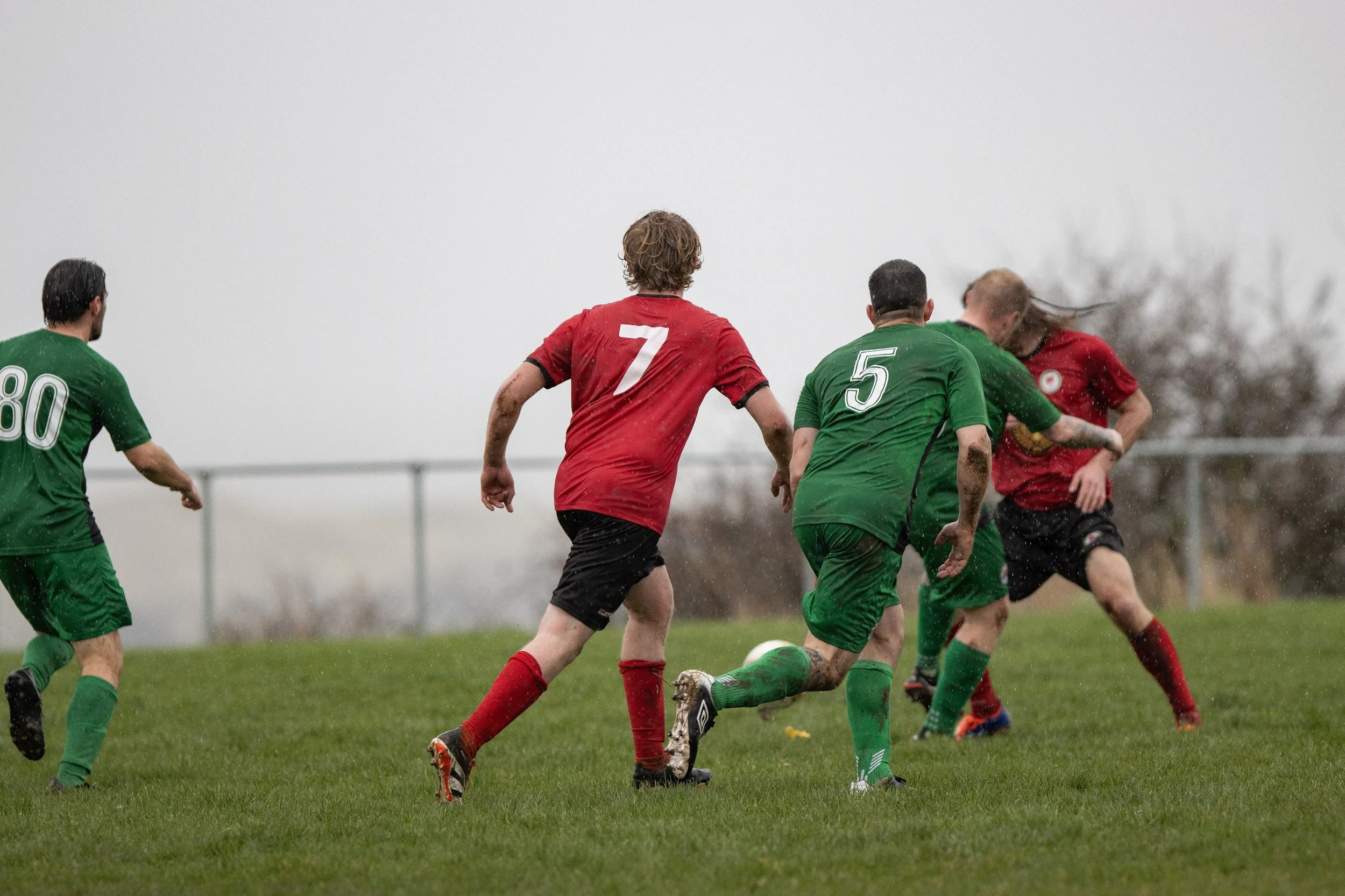 Soccer players on a field competing for the ball, some with muddy uniforms, under overcast weather.