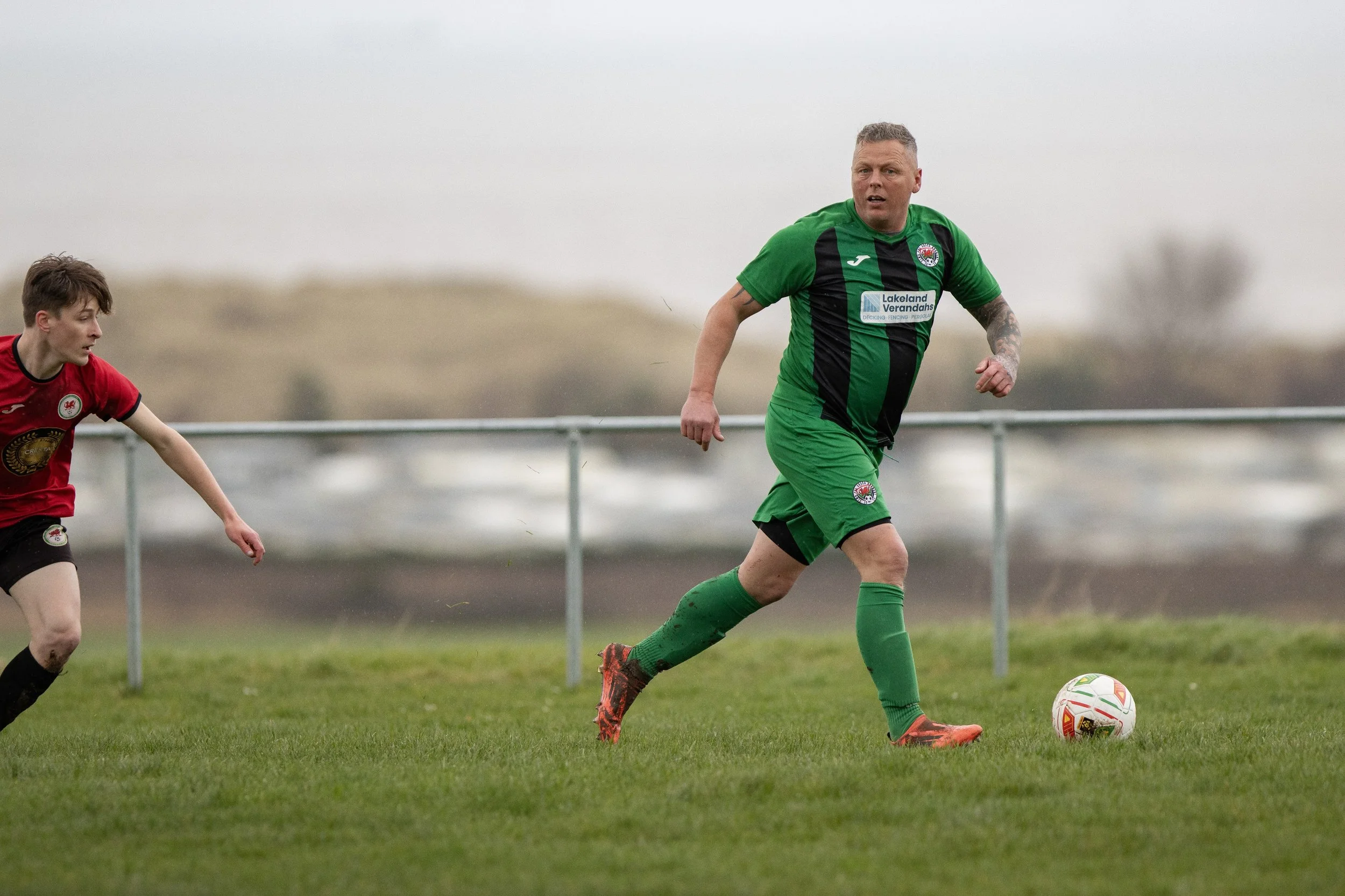 Two soccer players, one in a red jersey and one in a green and black jersey, compete for possession of the ball on an outdoor field with a blurred background and overcast sky.