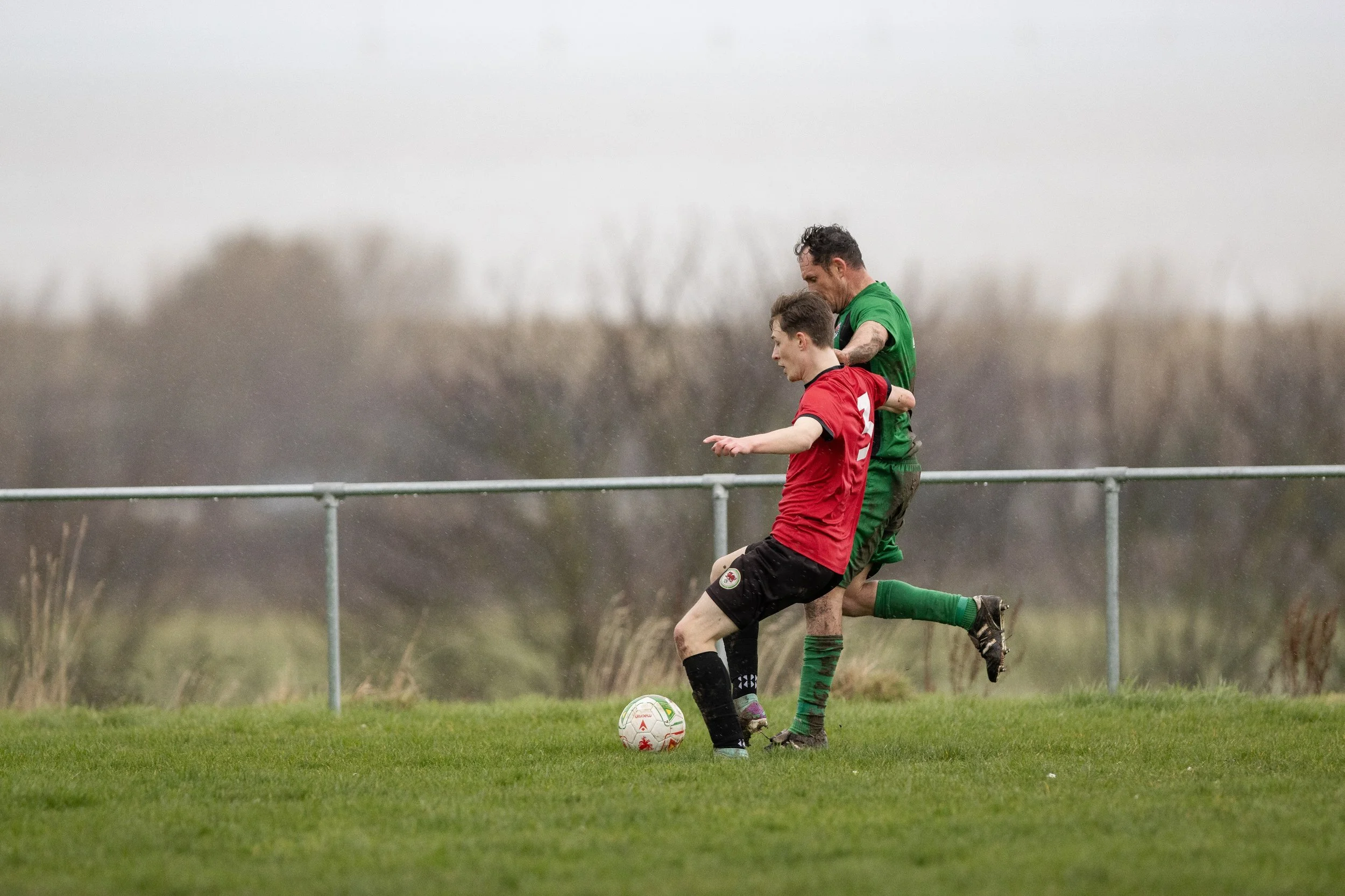 Two soccer players, one in a red jersey and the other in a green jersey, competing for the ball during a match on a grassy field under cloudy skies.
