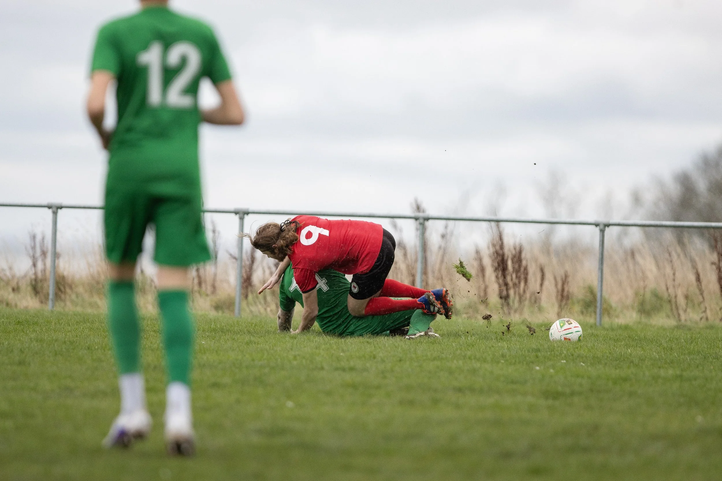 Two soccer players collide on the field while a third player in green stands nearby, with a soccer ball nearby. Overcast sky and grassy field in the background.