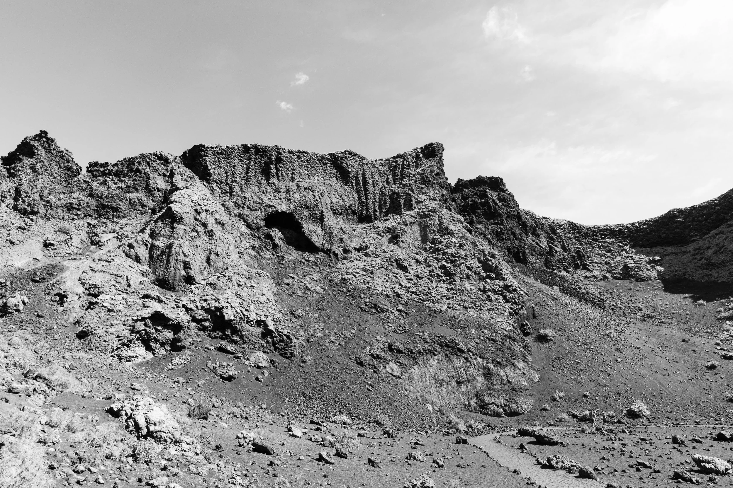 Black and white photograph of a rocky desert landscape with rugged cliffs, sparse vegetation, and a cloudy sky.