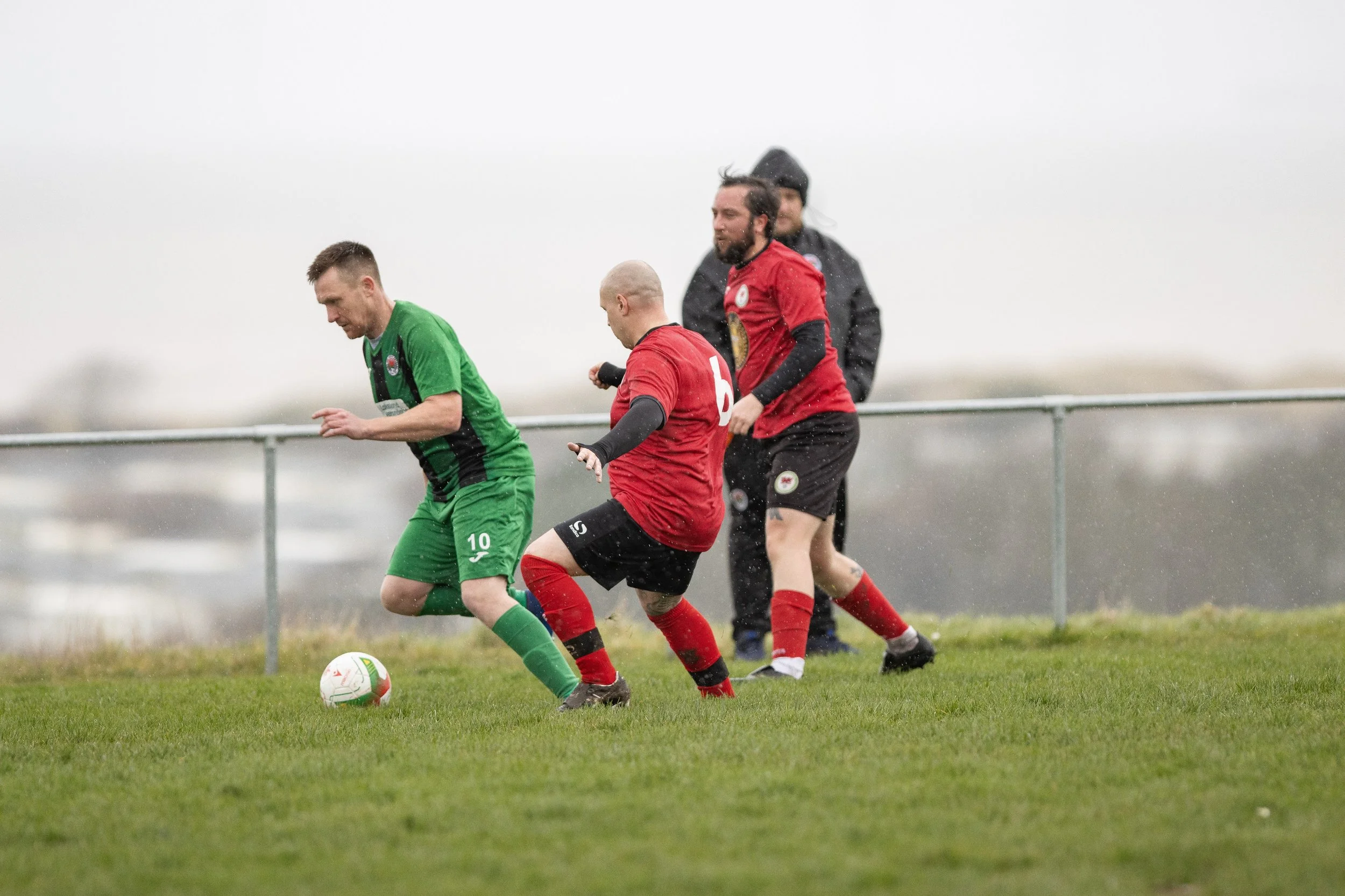 Soccer players in red and green jerseys playing on a grassy field under a gray sky, with a metal railing in the background.