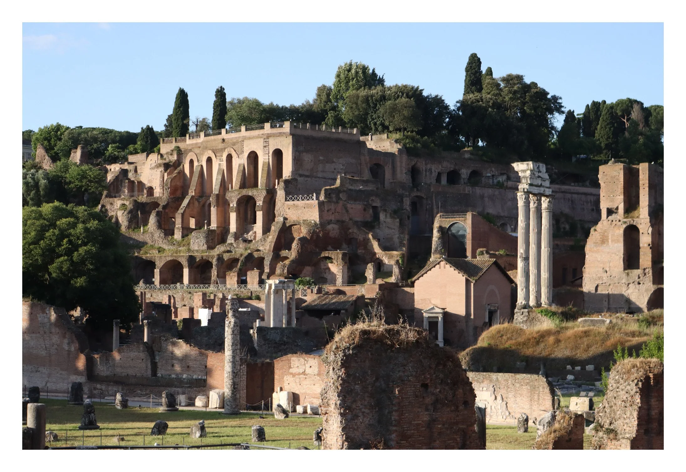 Ancient ruins with stone arches and columns on a hillside, surrounded by green trees and clear blue sky.