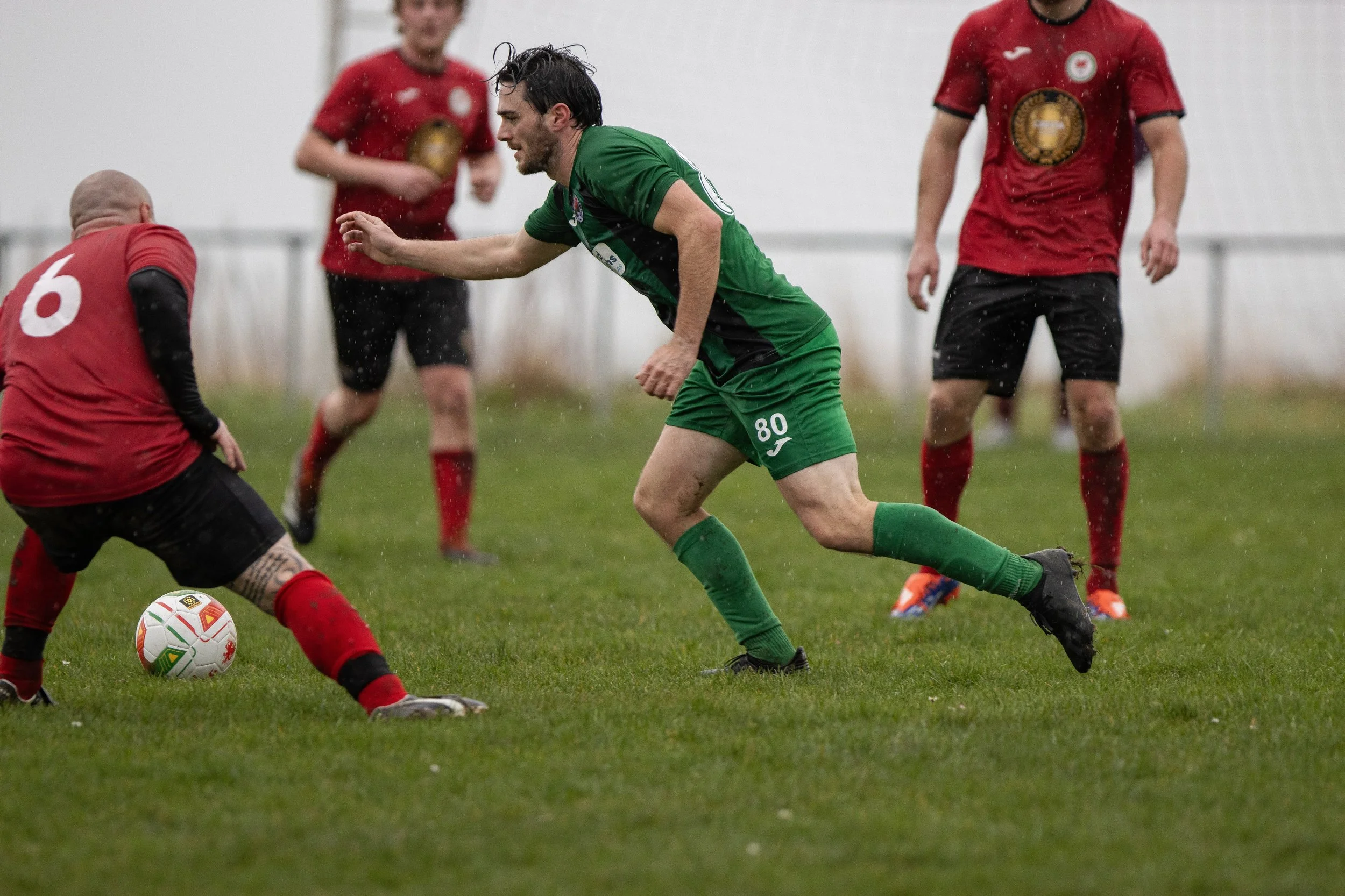 Soccer players competing for the ball on a rainy day, with one player in green and three players in red in the background.