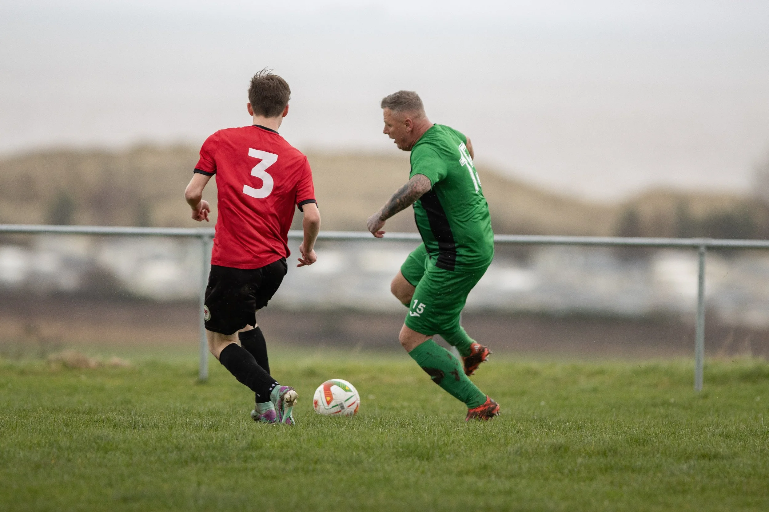 Two soccer players compete for the ball on the field, one in a red jersey with a large white number 3 on the back and the other in a green jersey with the number 15, during daytime with an overcast sky.