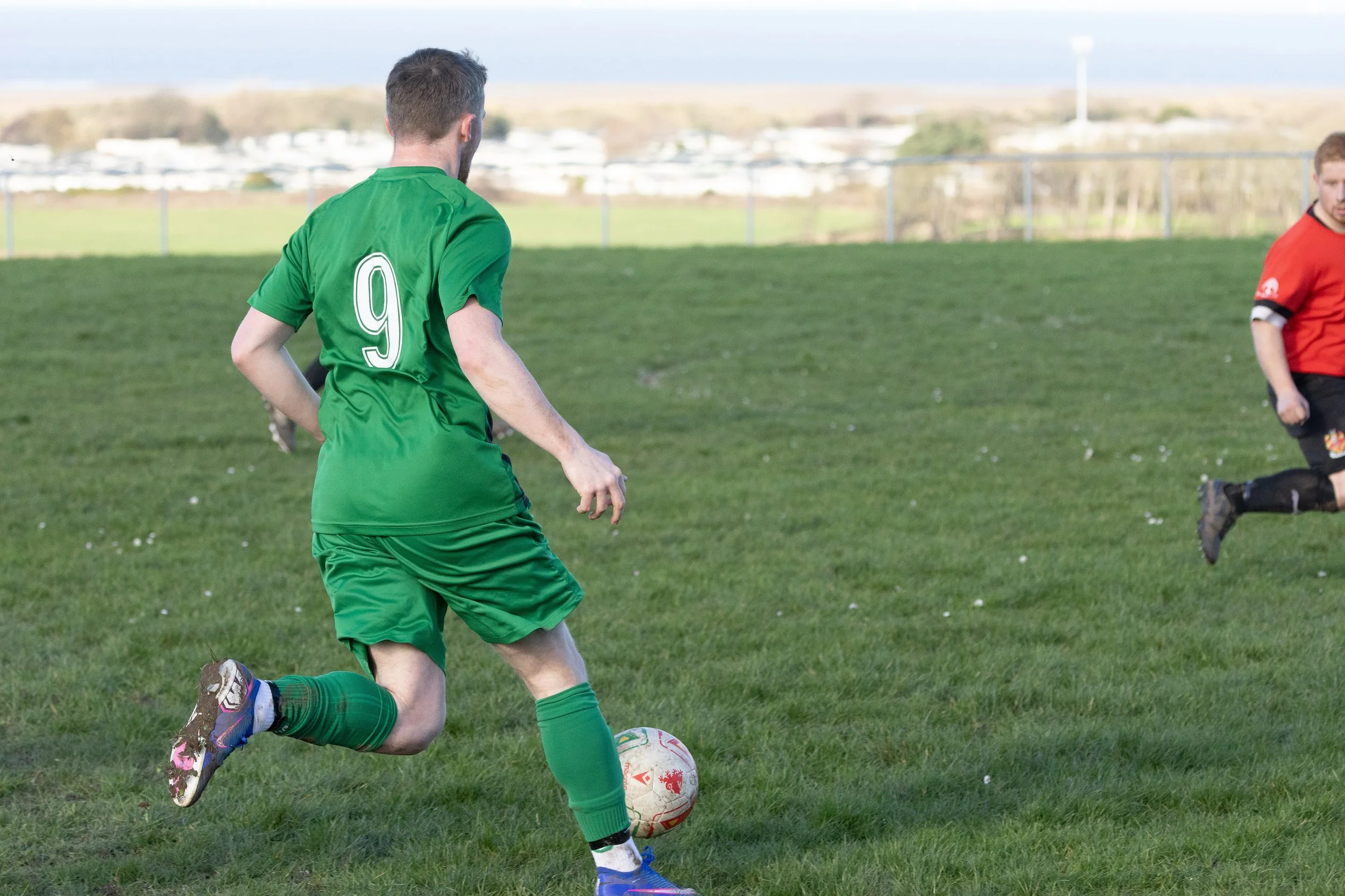 A soccer player in a green uniform with the number 9 on his back is kicking a soccer ball on a grassy field. Another player in a red and black uniform is visible in the background.
