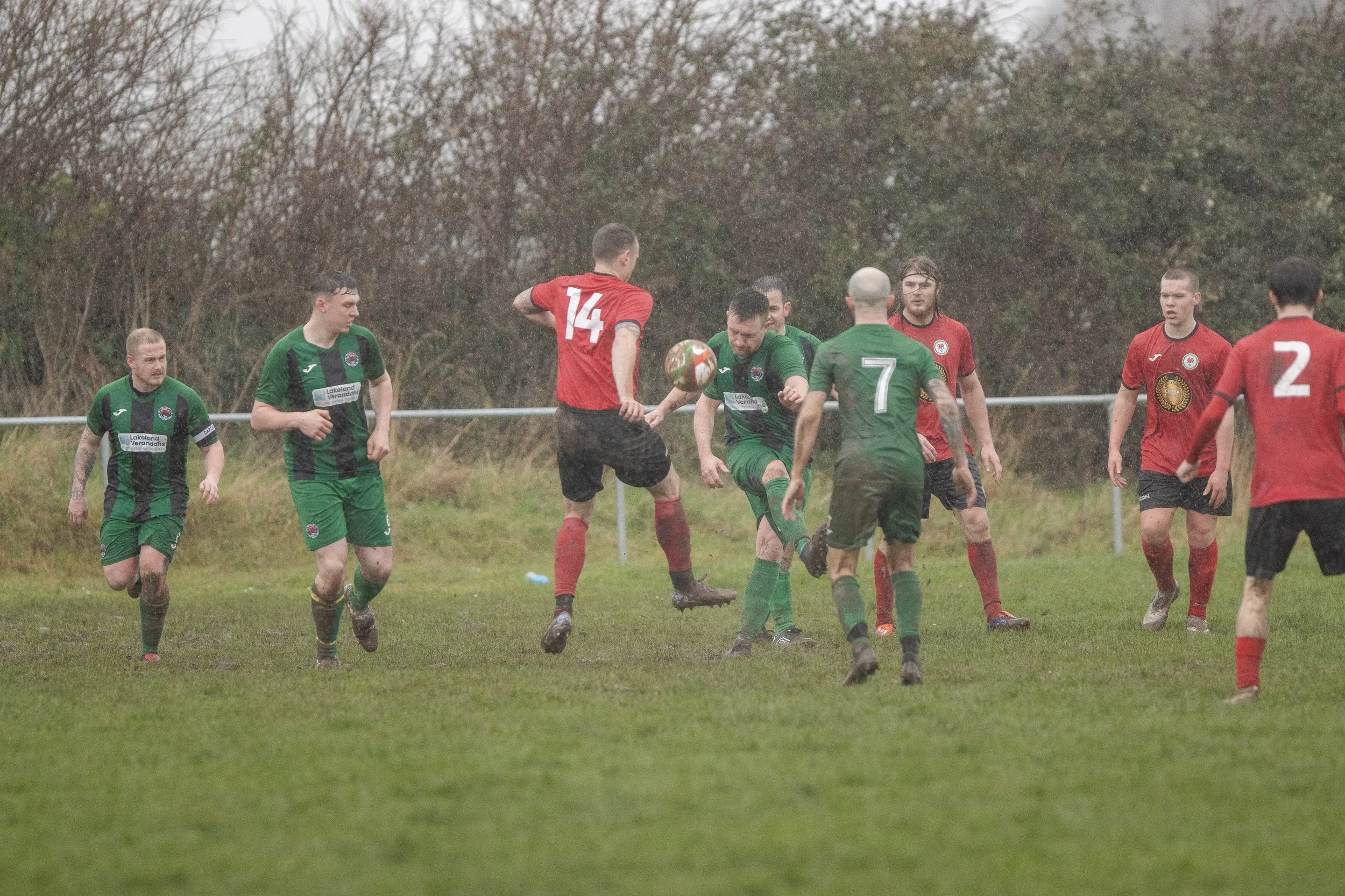Soccer players in green and red jerseys competing for the ball on a muddy field in rainy weather.
