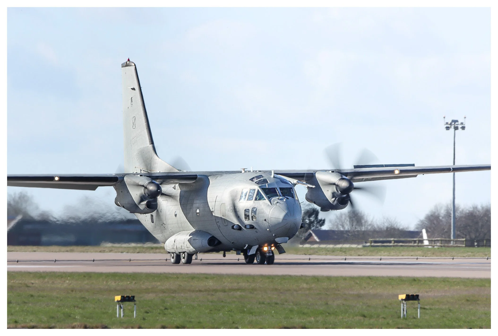 A military aircraft taxiing on the runway with two propellers spinning, against a partly cloudy sky and a weather tower in the background.