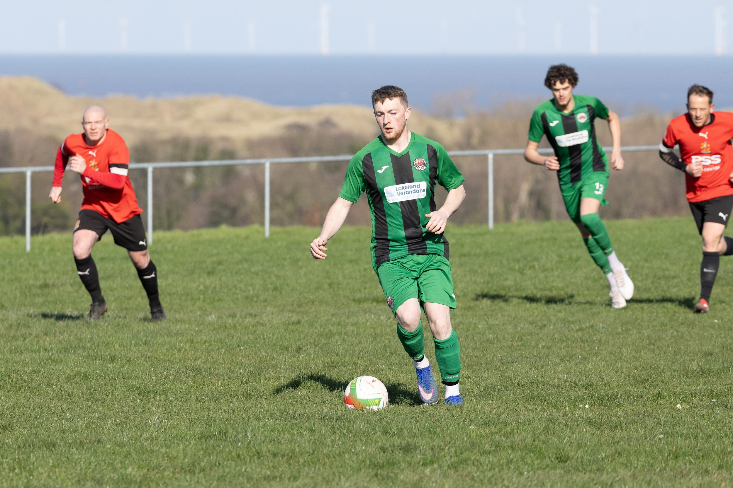 A soccer player in a green and black uniform is dribbling a soccer ball on a grassy field, with three opponents in red and black uniforms running behind him, during daytime with wind turbines visible in the background.
