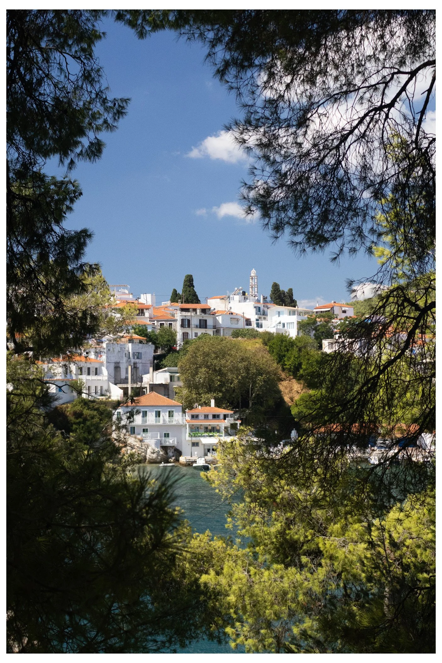 Scenic view of a hillside town with white houses and red-tiled roofs, surrounded by green trees, overlooking water, under a blue sky with white clouds.