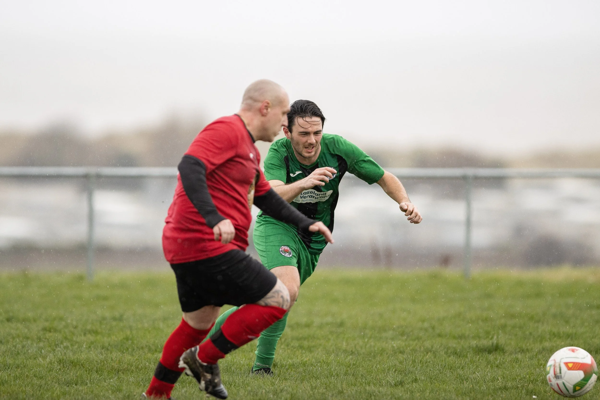 Two male soccer players compete for the ball on a rainy day on a grassy field.