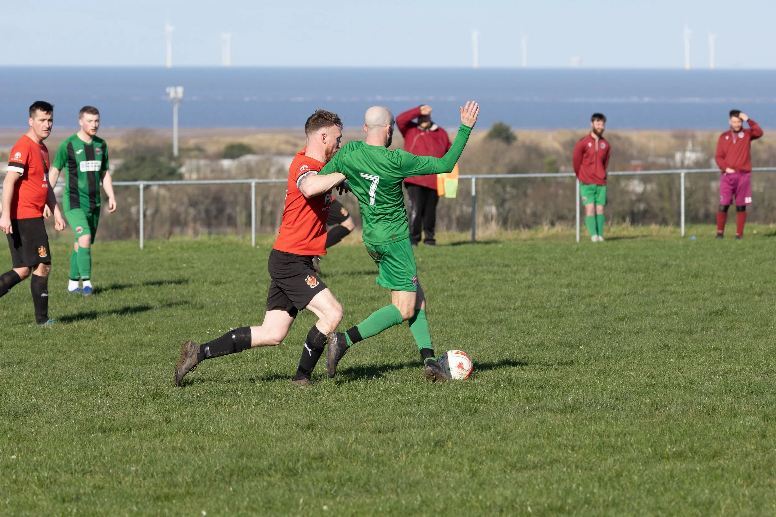 Two soccer players compete for the ball on a grassy field, with additional players and spectators in the background, near a fence and wind turbines beyond.