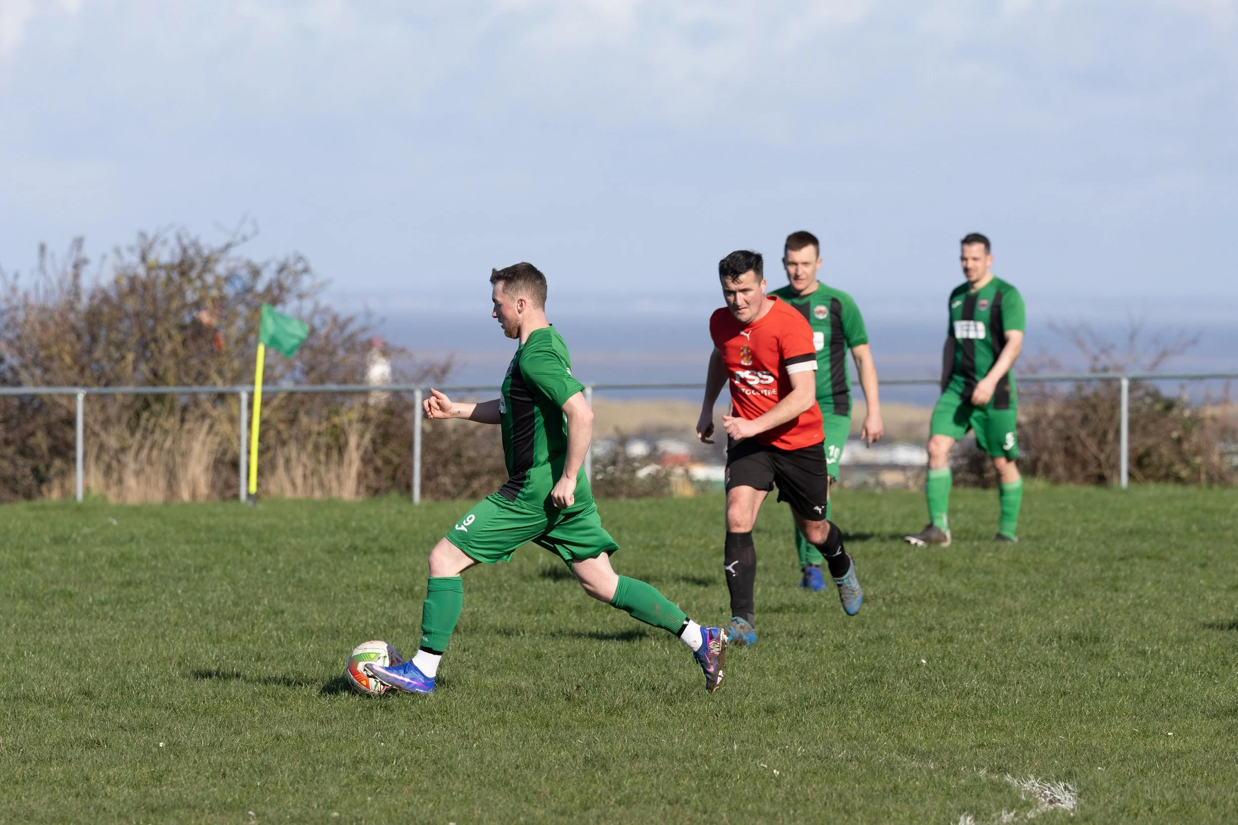 Four soccer players, three in green jerseys and one in a red and black jersey, are on a grassy field during a match. One player in green is about to kick the soccer ball, while the other players are jogging or standing nearby. A yellow corner flag is