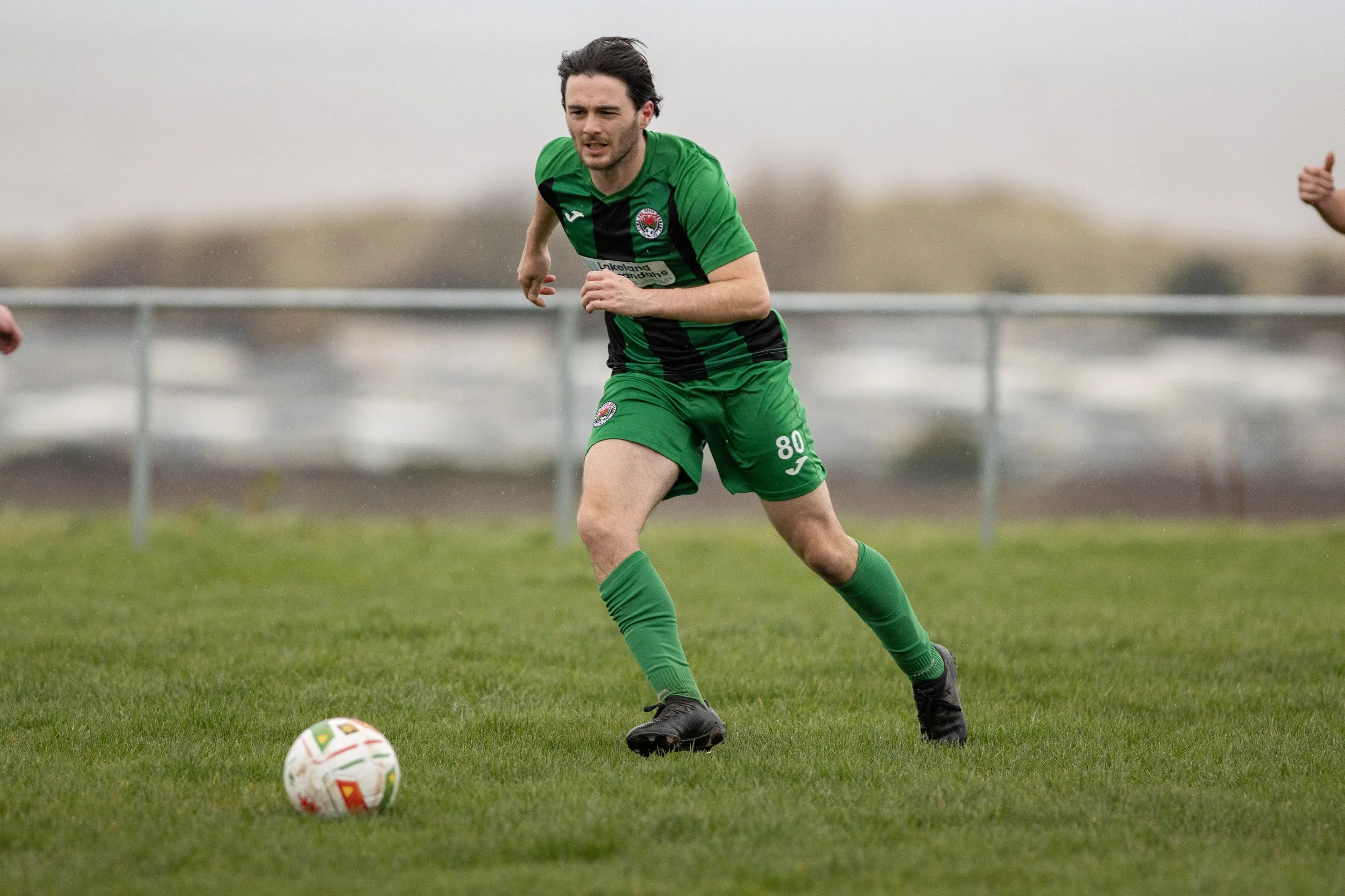 A soccer player in a green uniform is running on the field, approaching a soccer ball. The background shows a metal fence and overcast sky.