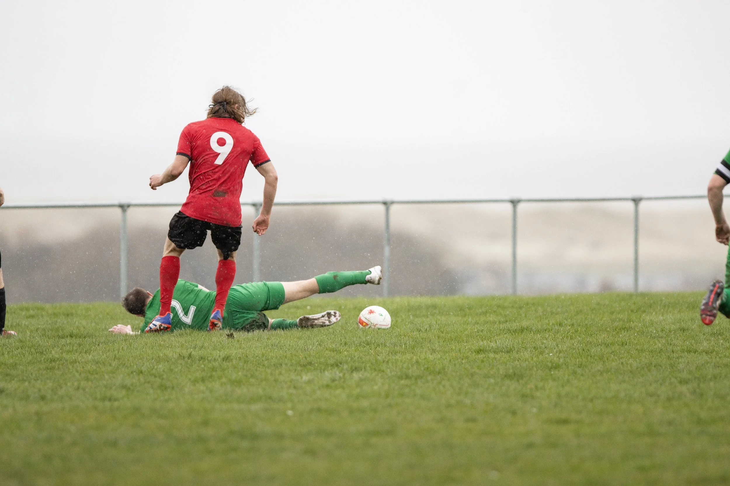 Soccer game with a player in a red jersey and black shorts about to score on a goalkeeper in green who is on the ground.