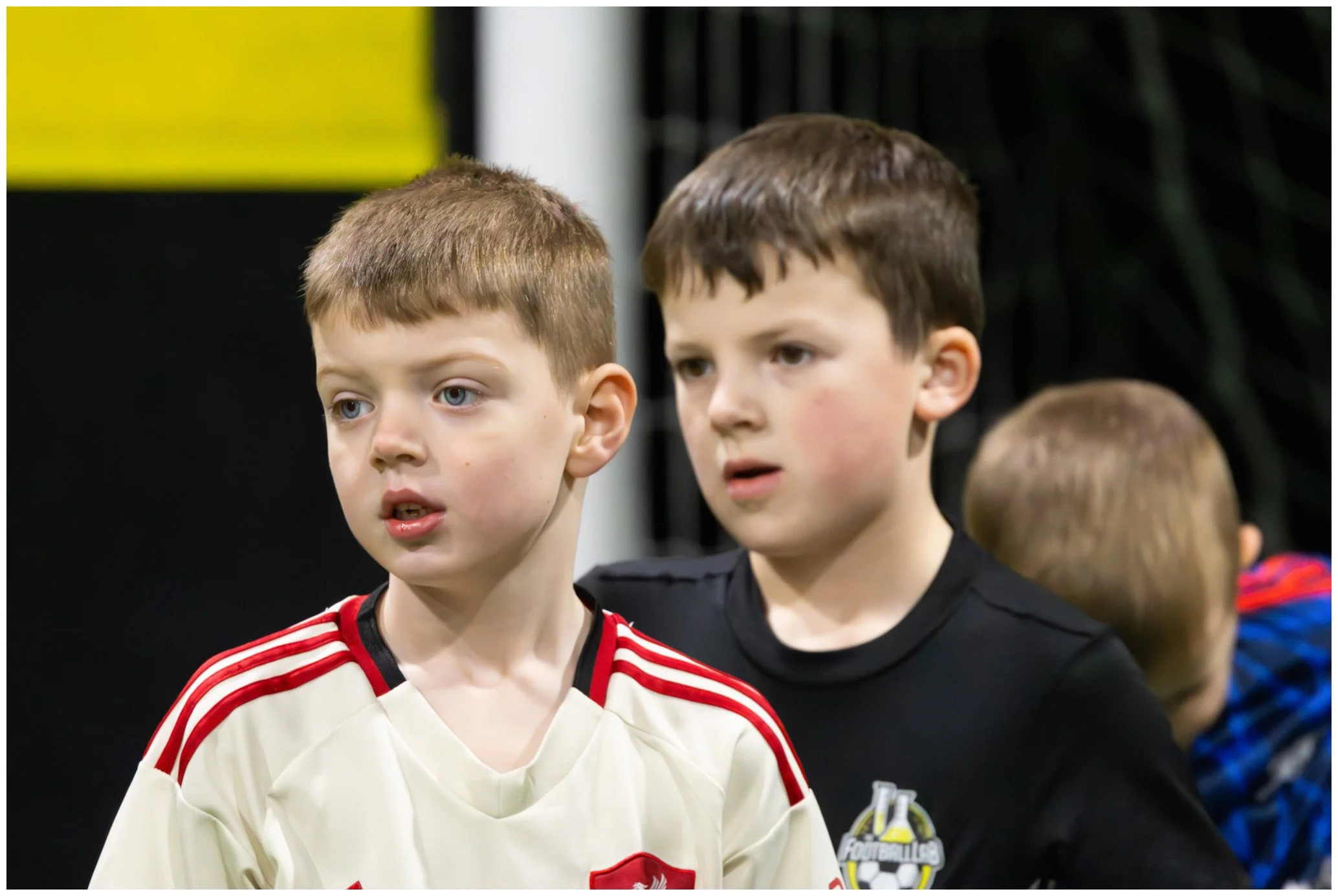 Two young boys with serious expressions, standing close together indoors, one wearing a white sports jersey with red accents, the other in a black shirt with a logo, behind a faintly visible third child in a blue jacket.