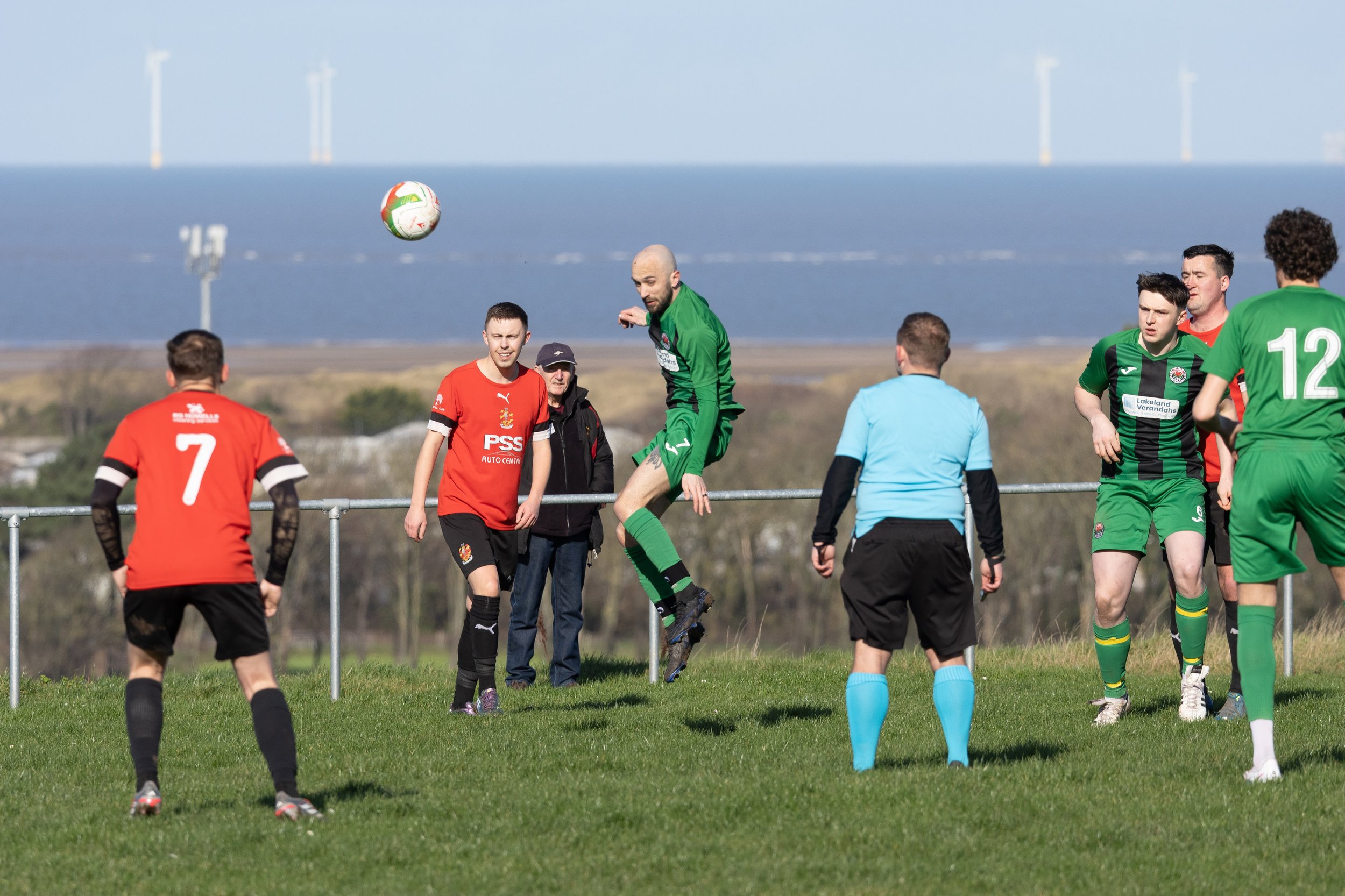 A soccer match with players in red and green jerseys, a referee, and a player jumping to head the ball near the goal, with a landscape and wind turbines in the background.