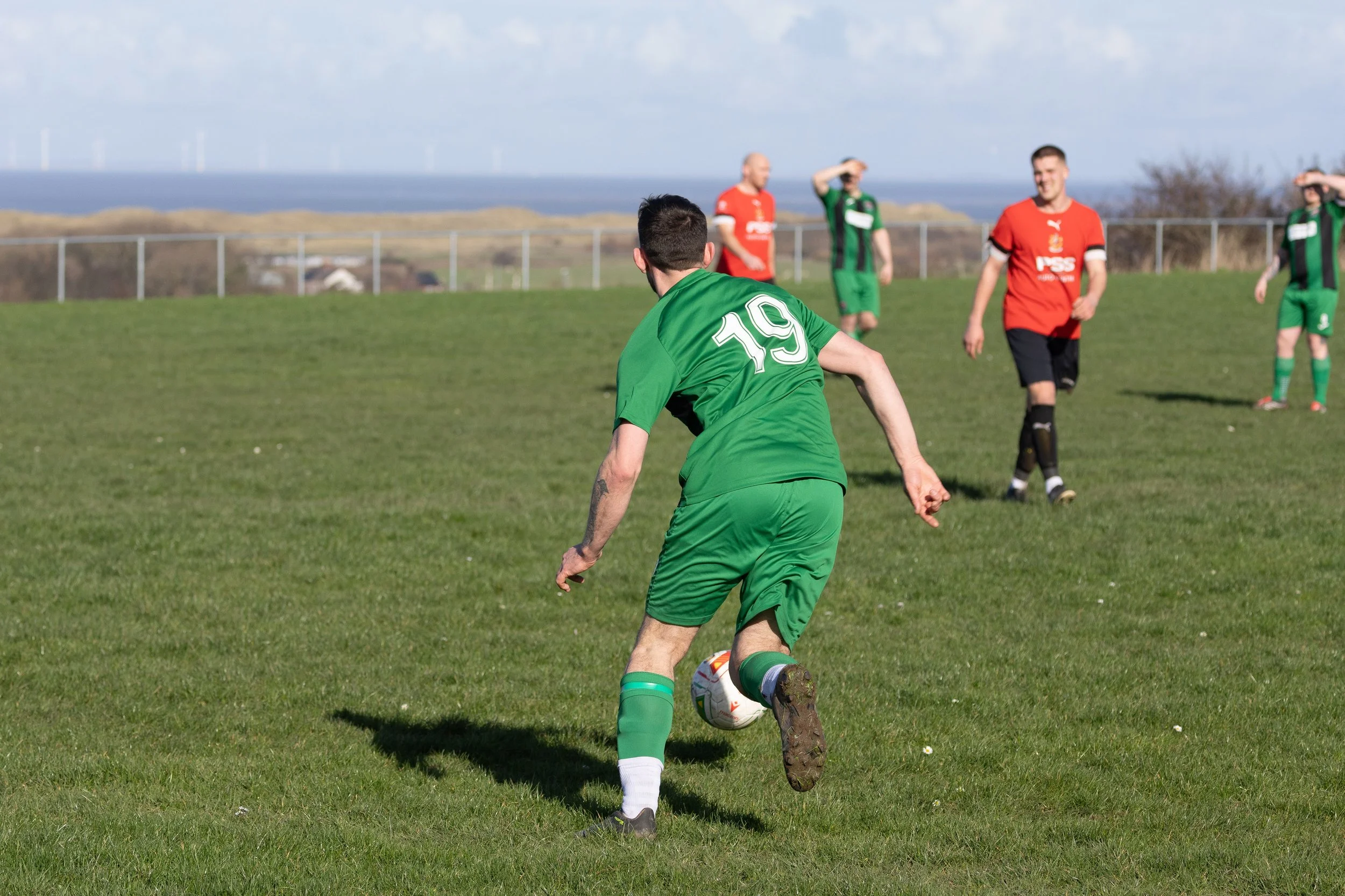 A soccer player wearing a green jersey with the number 19 is kicking a ball on a grassy field. Other players in red and green jerseys are visible in the background, with a fence and a landscape with wind turbines and water in the distance.