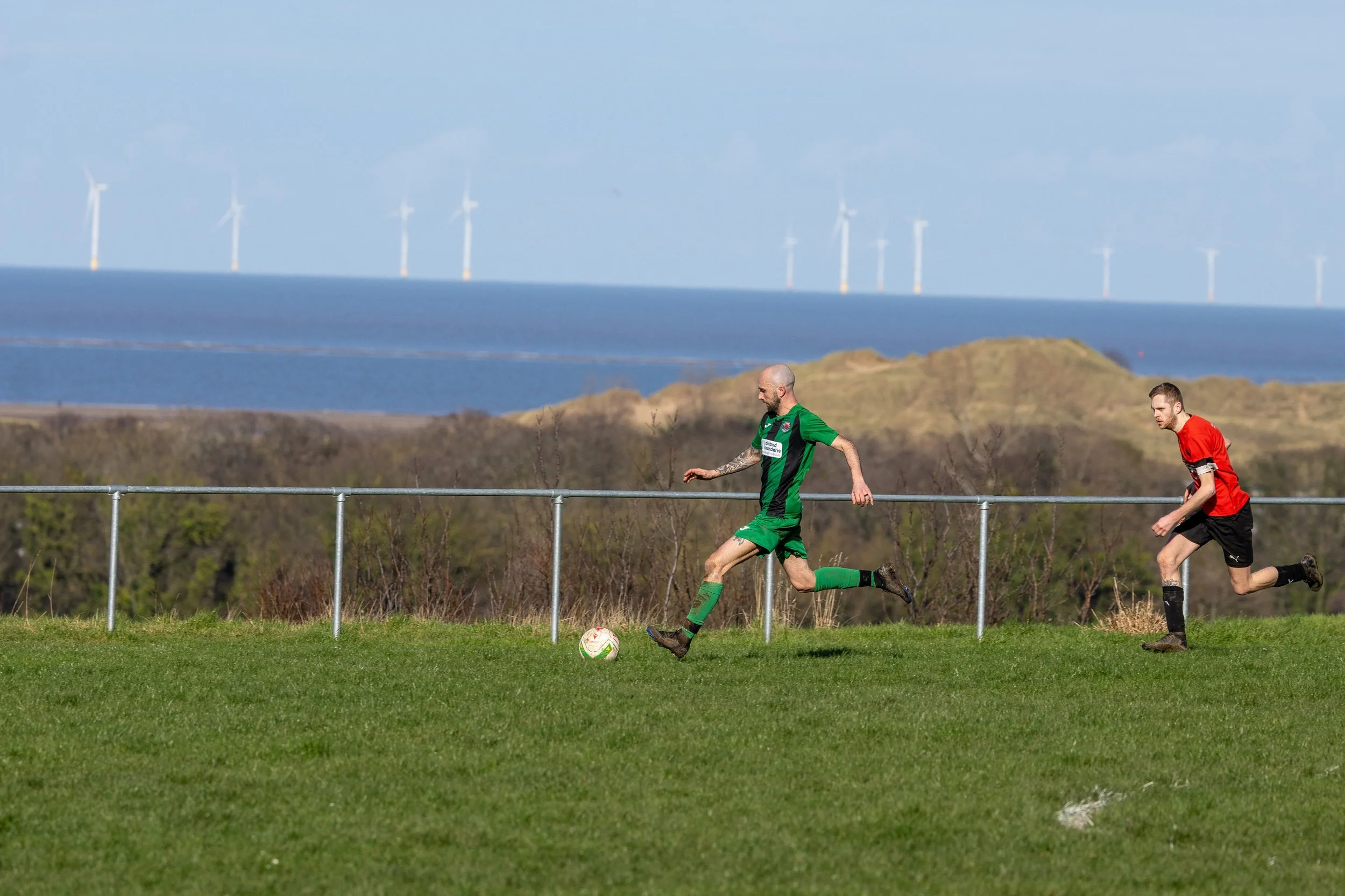 Two soccer players on a grassy field with a scenic background of hills, water, and wind turbines, one in a green uniform kicking a soccer ball, the other in a red uniform running behind.