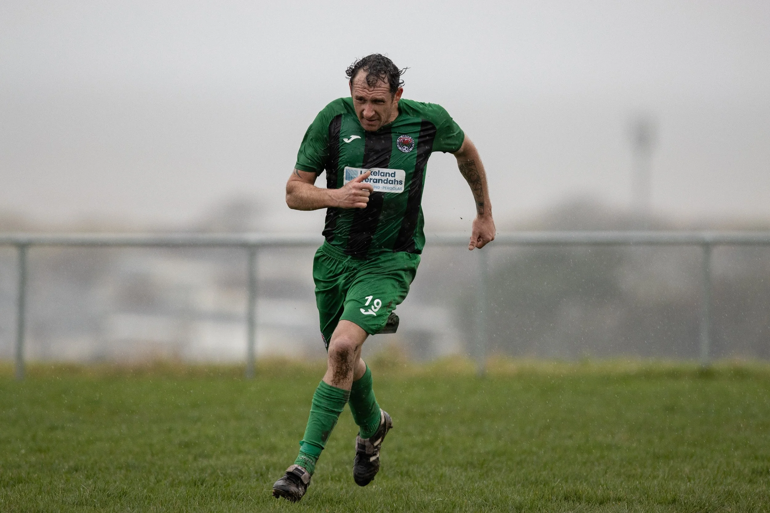 A man in a green and black soccer uniform running on a grass field during a rainy day.