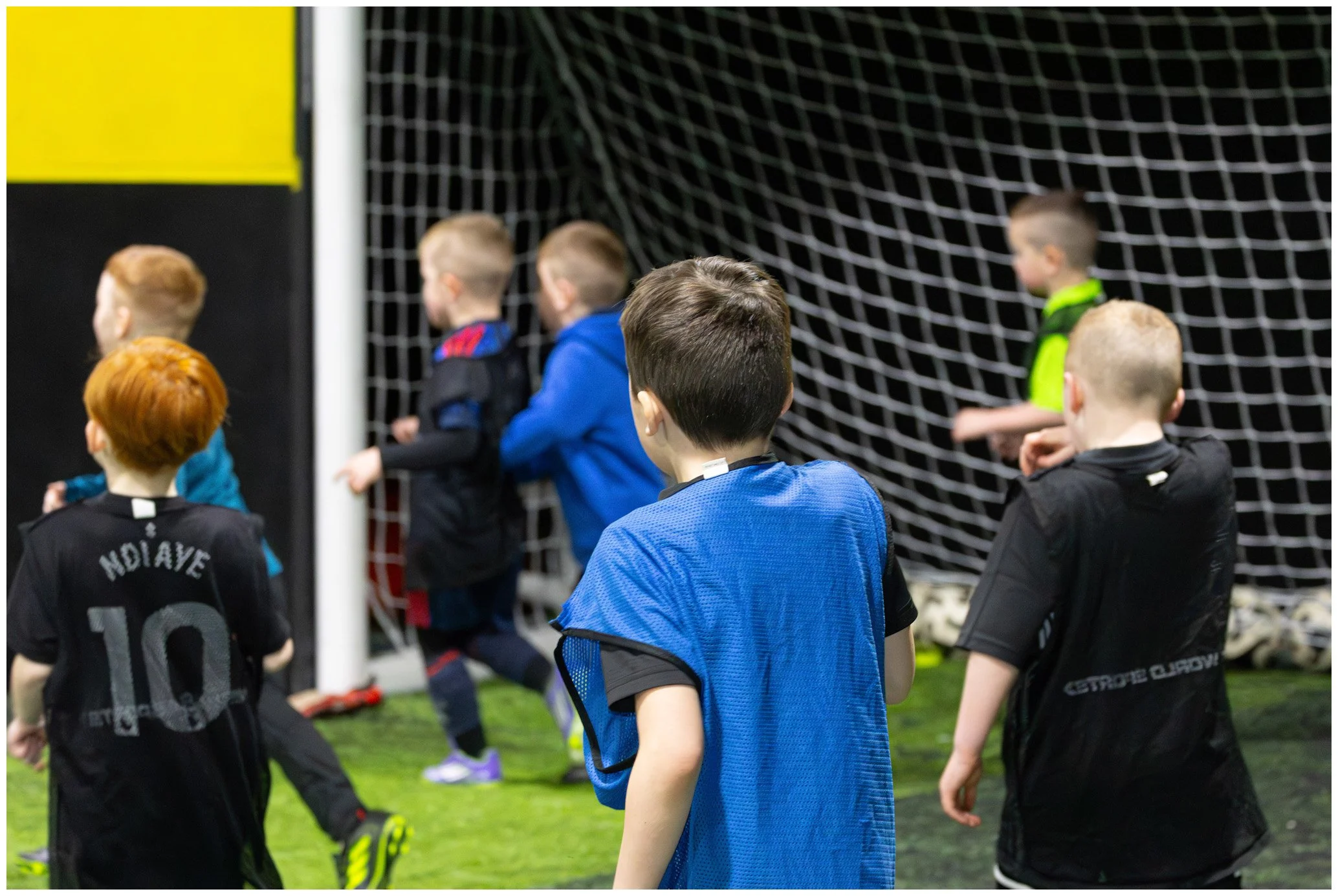 Group of young children at a soccer practice or game, standing near the goal net on indoor turf field.