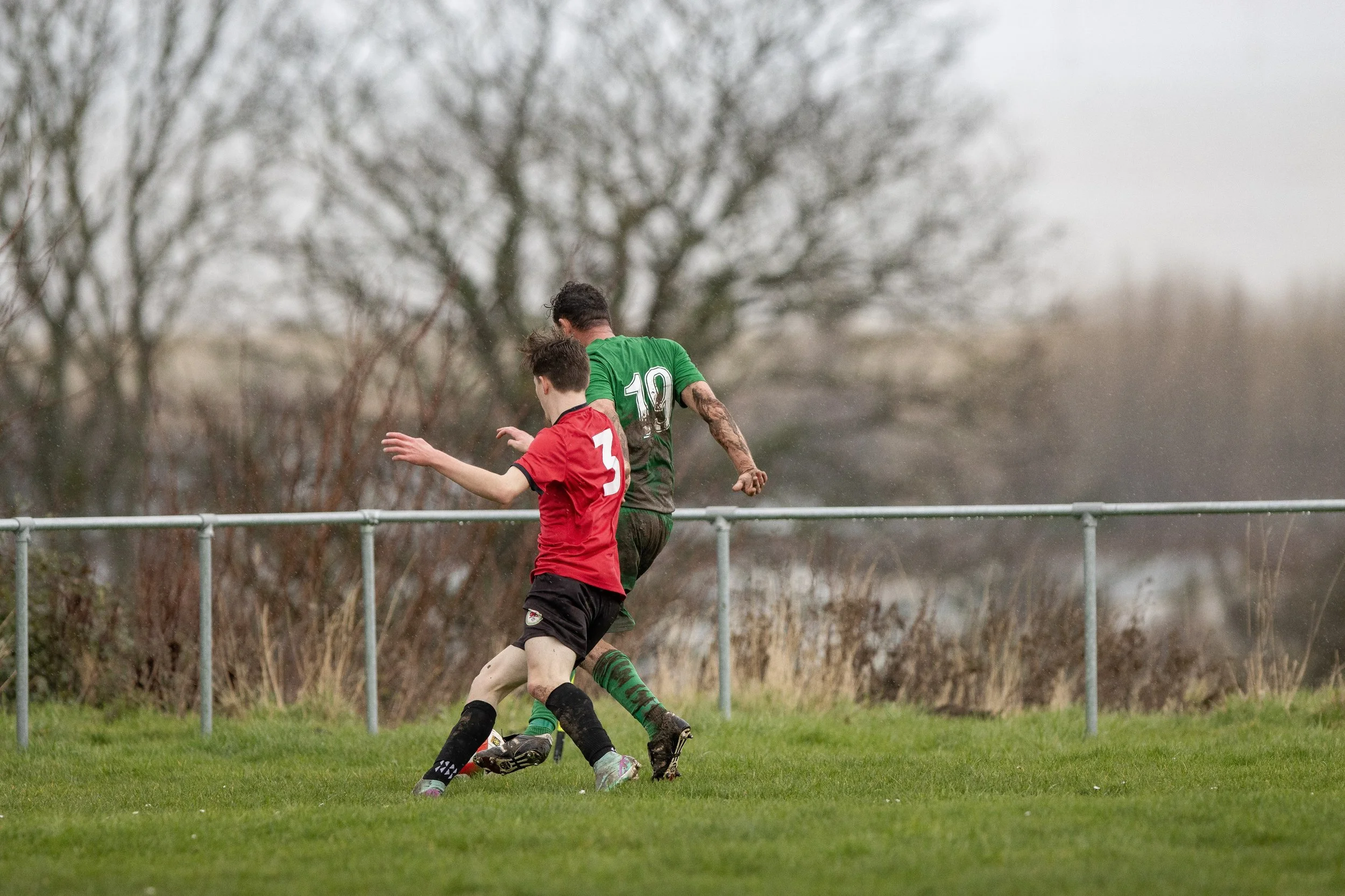 Two soccer players are competing for the ball on a grassy field, with a metal fence and leafless trees in the background during overcast weather.