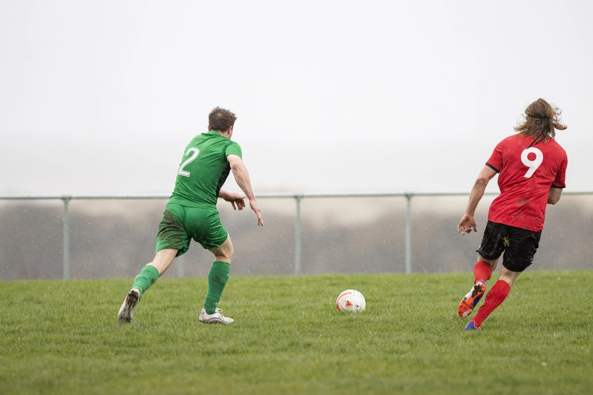 Two soccer players, one in green jersey number 2, the other in red jersey number 9, chase the soccer ball on a grassy field on a cloudy day.