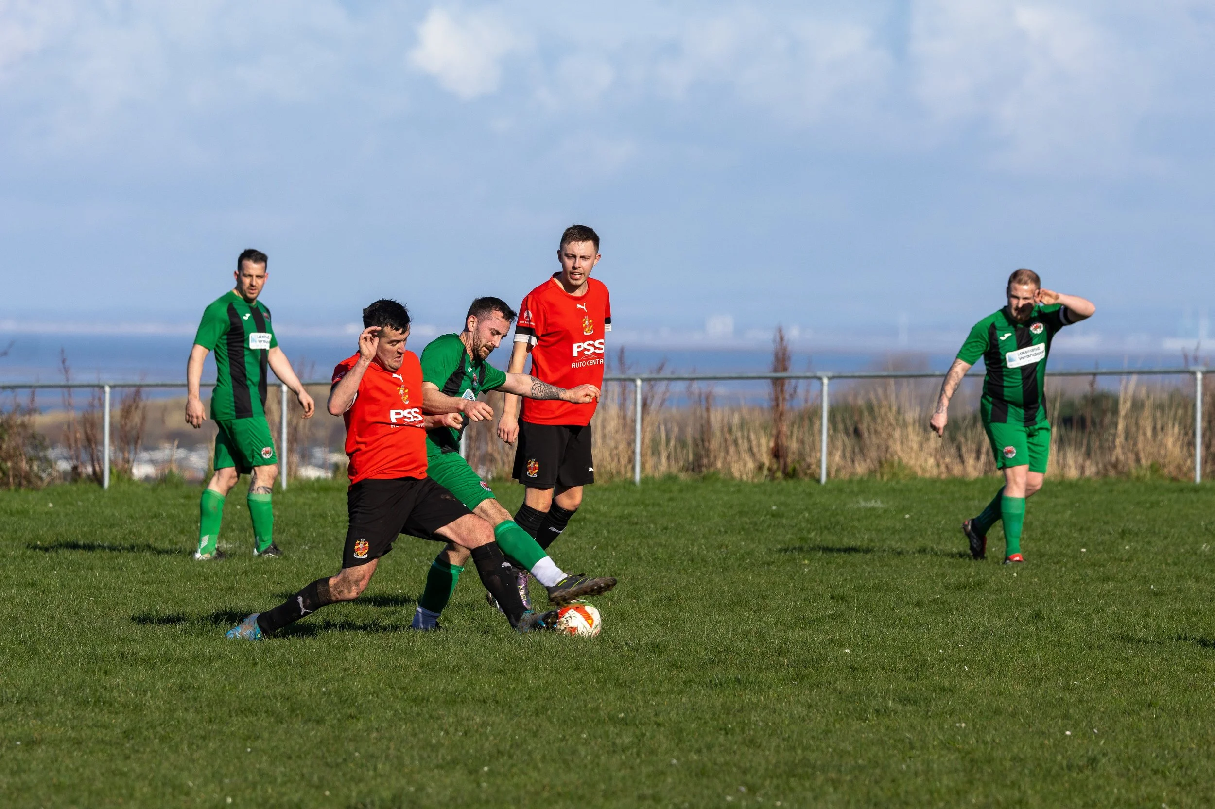Soccer players in green and red uniforms playing on a grassy field with a fence and cloudy sky in the background.