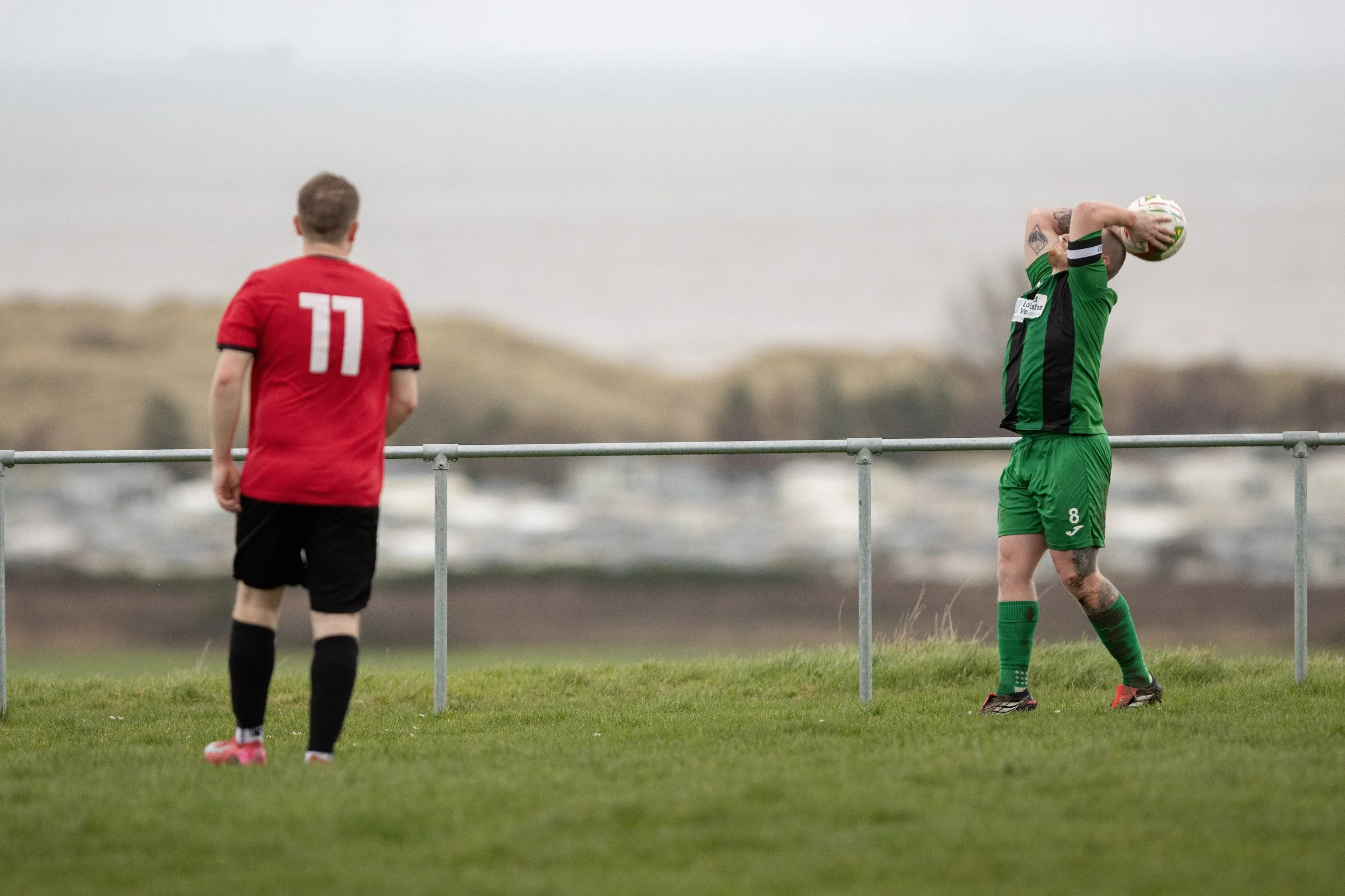 A soccer player wearing a green uniform prepares to take a free kick, while another player in a red uniform stands nearby on a grassy field.