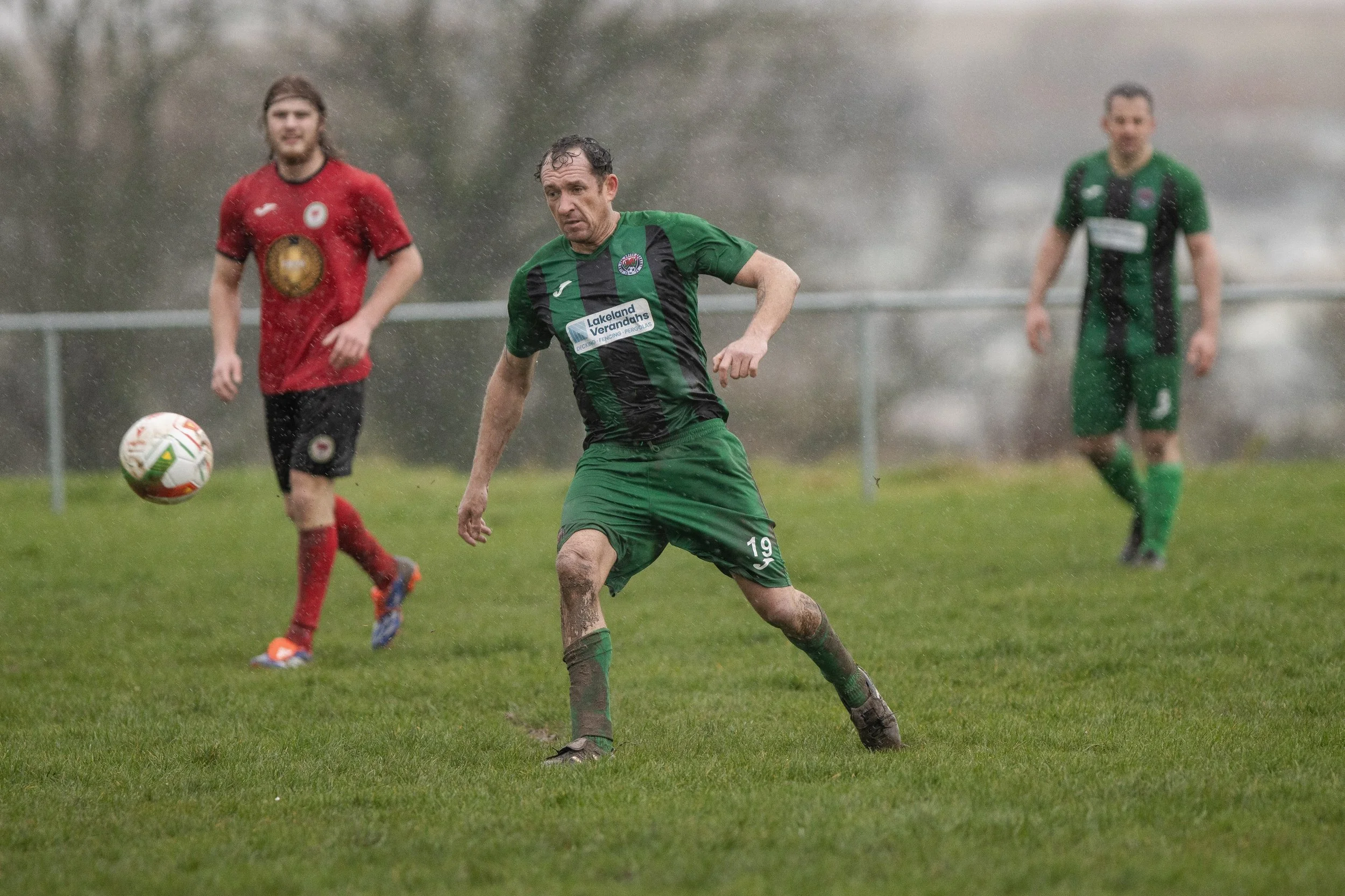 Soccer players playing in the rain on a grassy field, with one player in green and black approaching the ball while two players in red and green watch in the background.