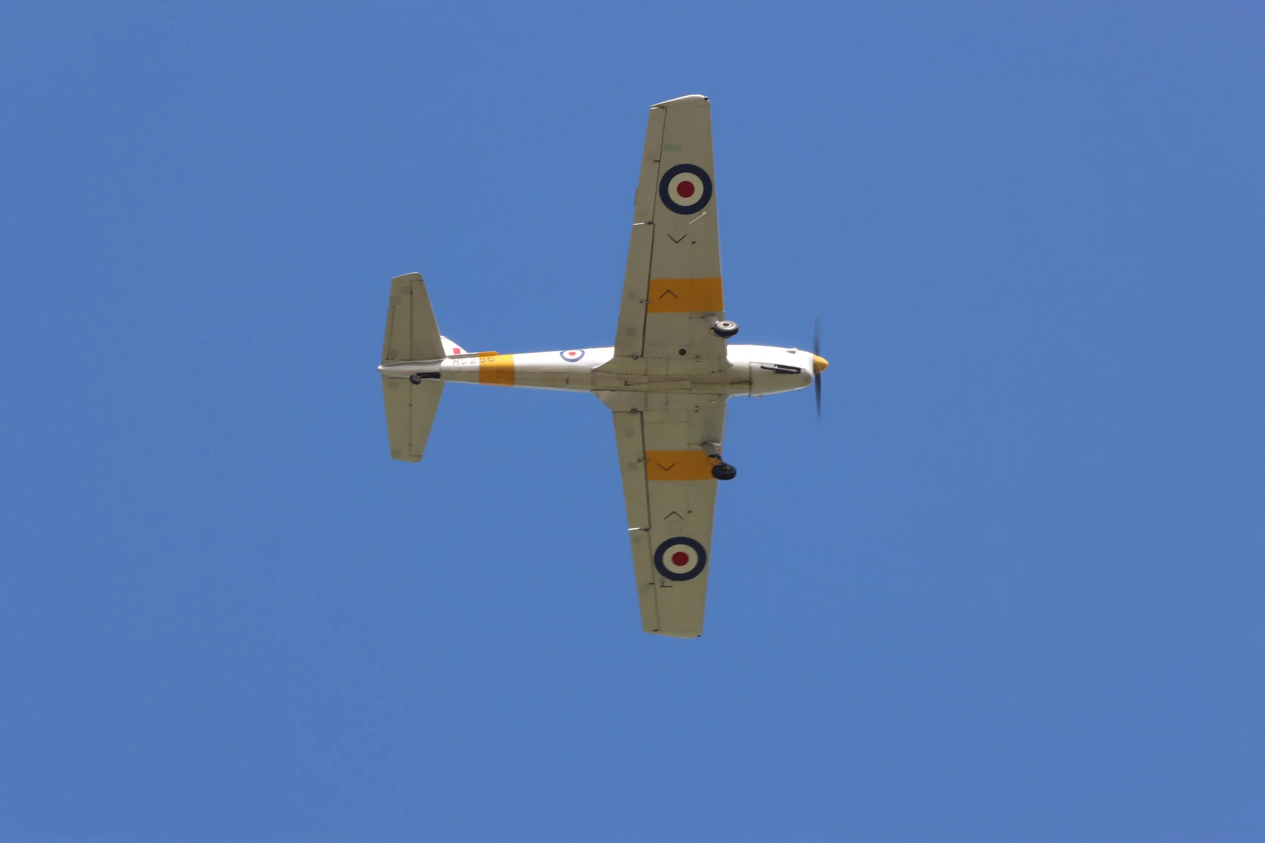 A white aircraft with roundels and yellow markings flying against a clear blue sky.