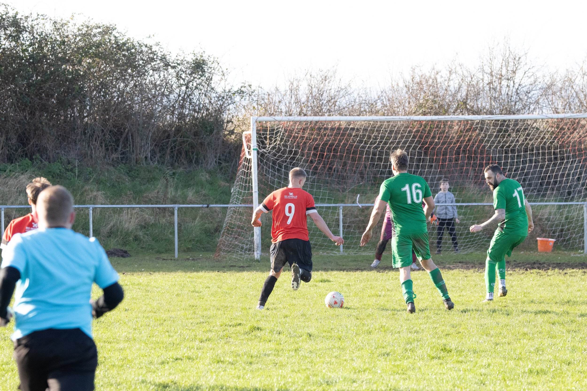 Soccer players in green and red uniforms compete near the goal, with one player in red about to kick the ball while others prepare to defend, on a grassy field with trees in the background.