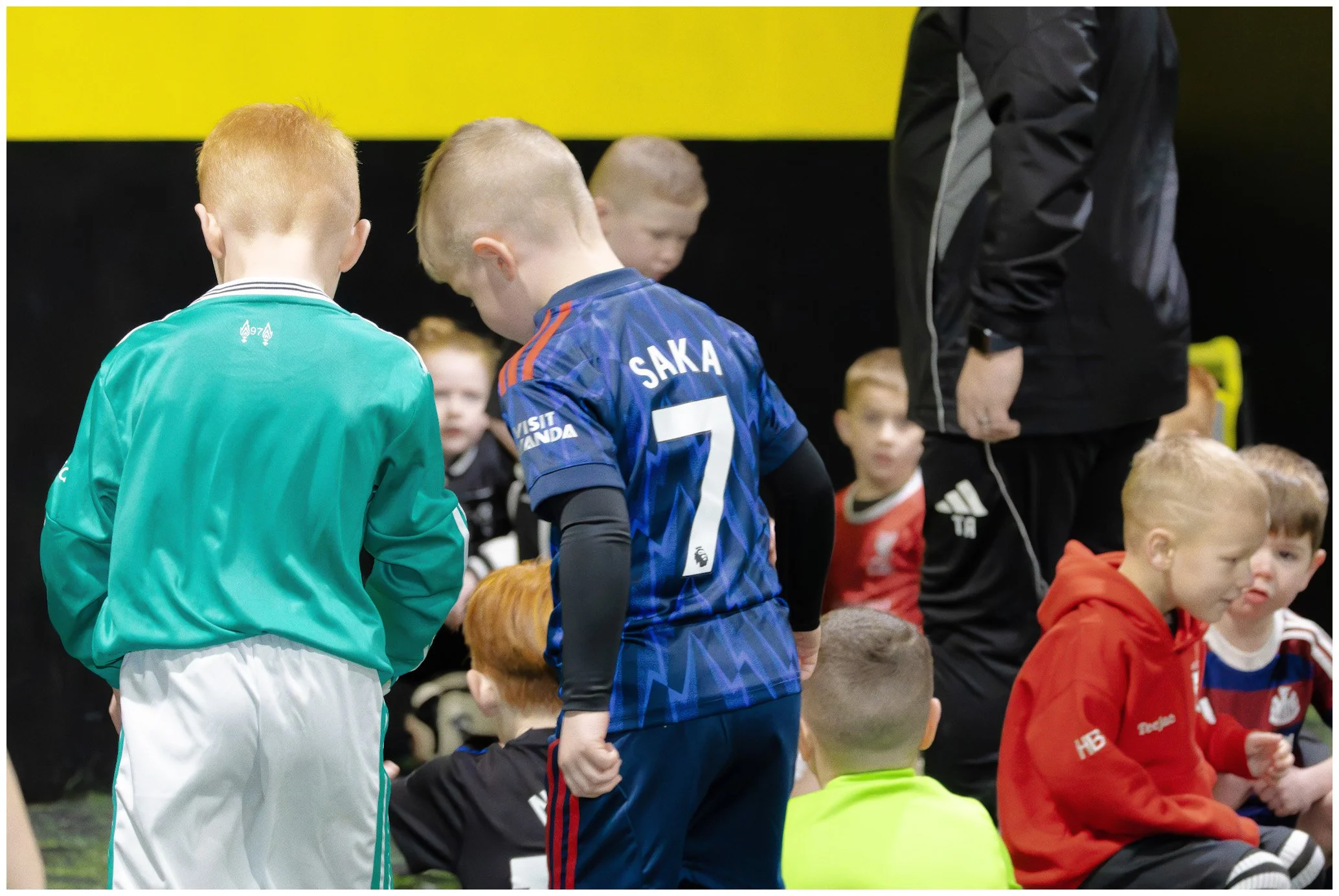 Group of children and a coach in a sports setting. Children are wearing sports jerseys and sitting on the ground. One child in a blue jersey with the name Saka and number 7 on the back is standing and talking to another child in a green jacket.