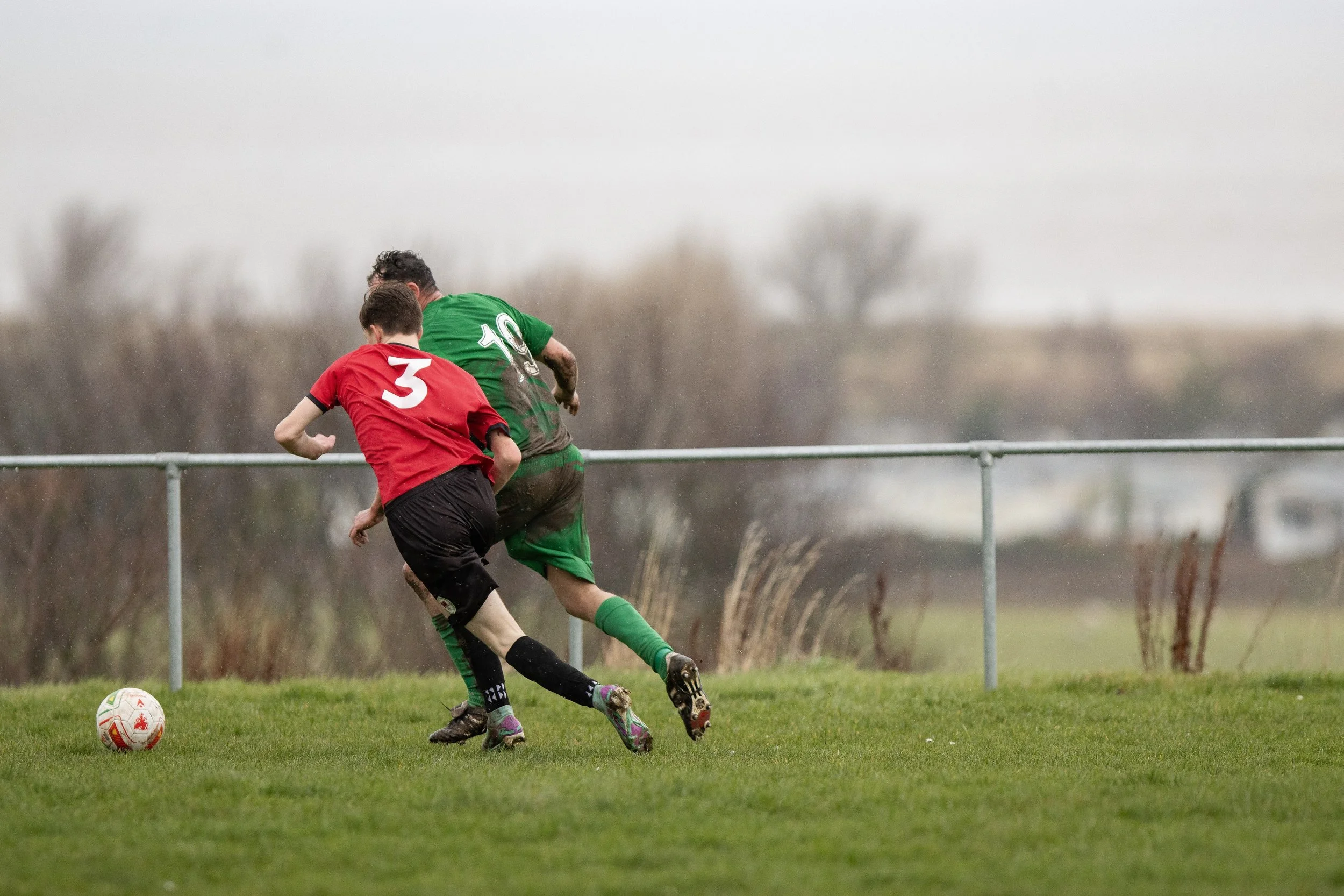 Two soccer players, one in red and one in green, battling for the ball on a grassy field