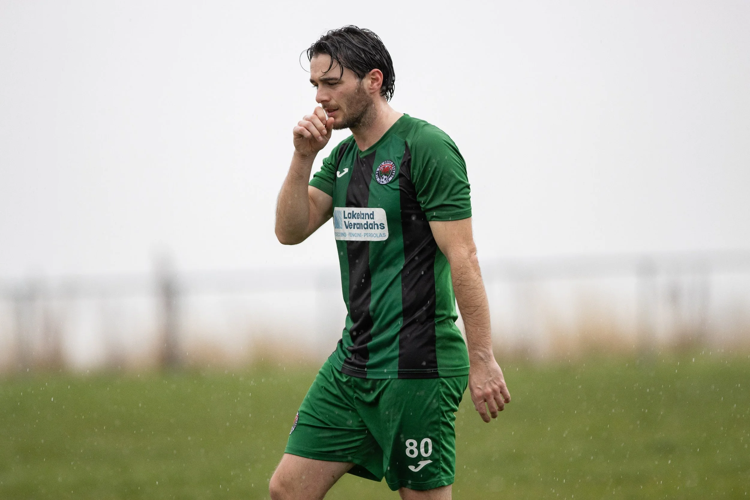 Soccer player in green and black uniform with number 80, standing on field with rain, hand near mouth.