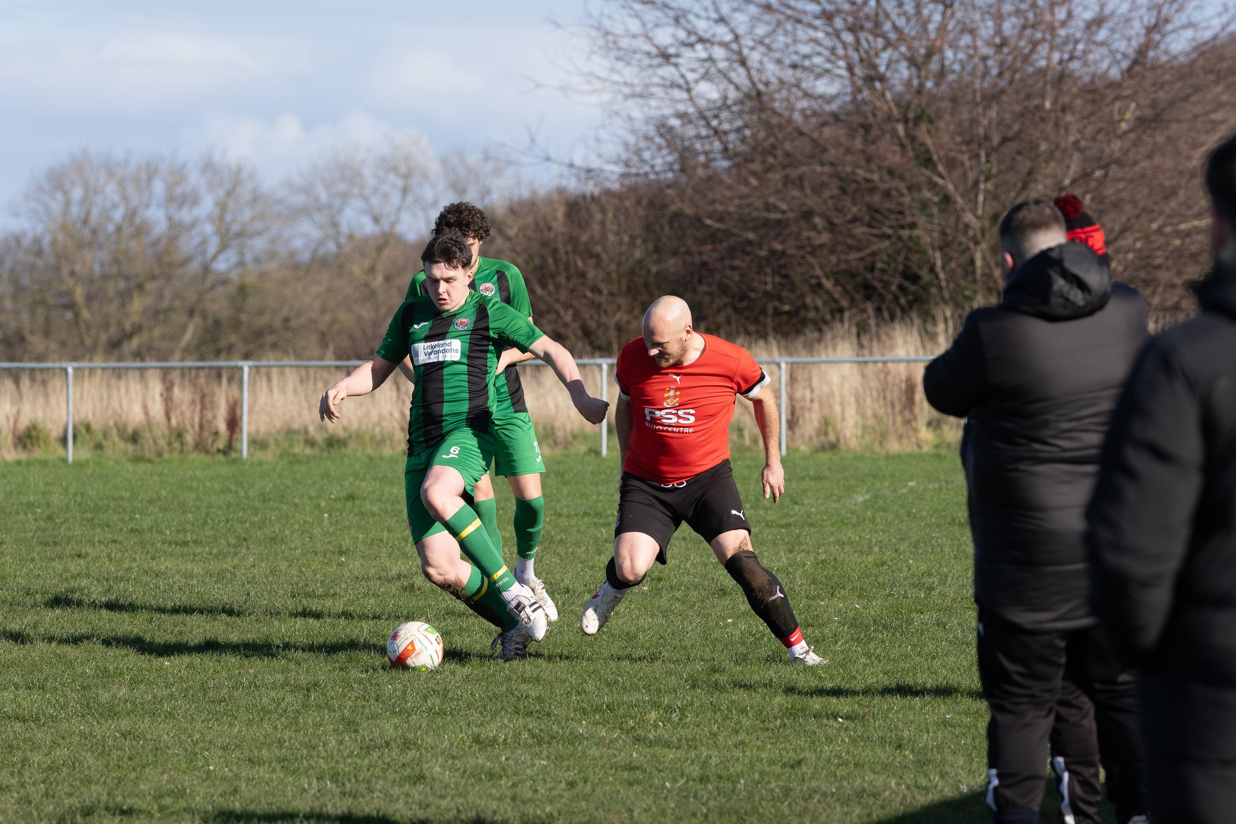 Two soccer players, one in a green and black uniform and another in a red and black uniform, competing for the ball on a grassy field. Two spectators are watching from the sidelines.