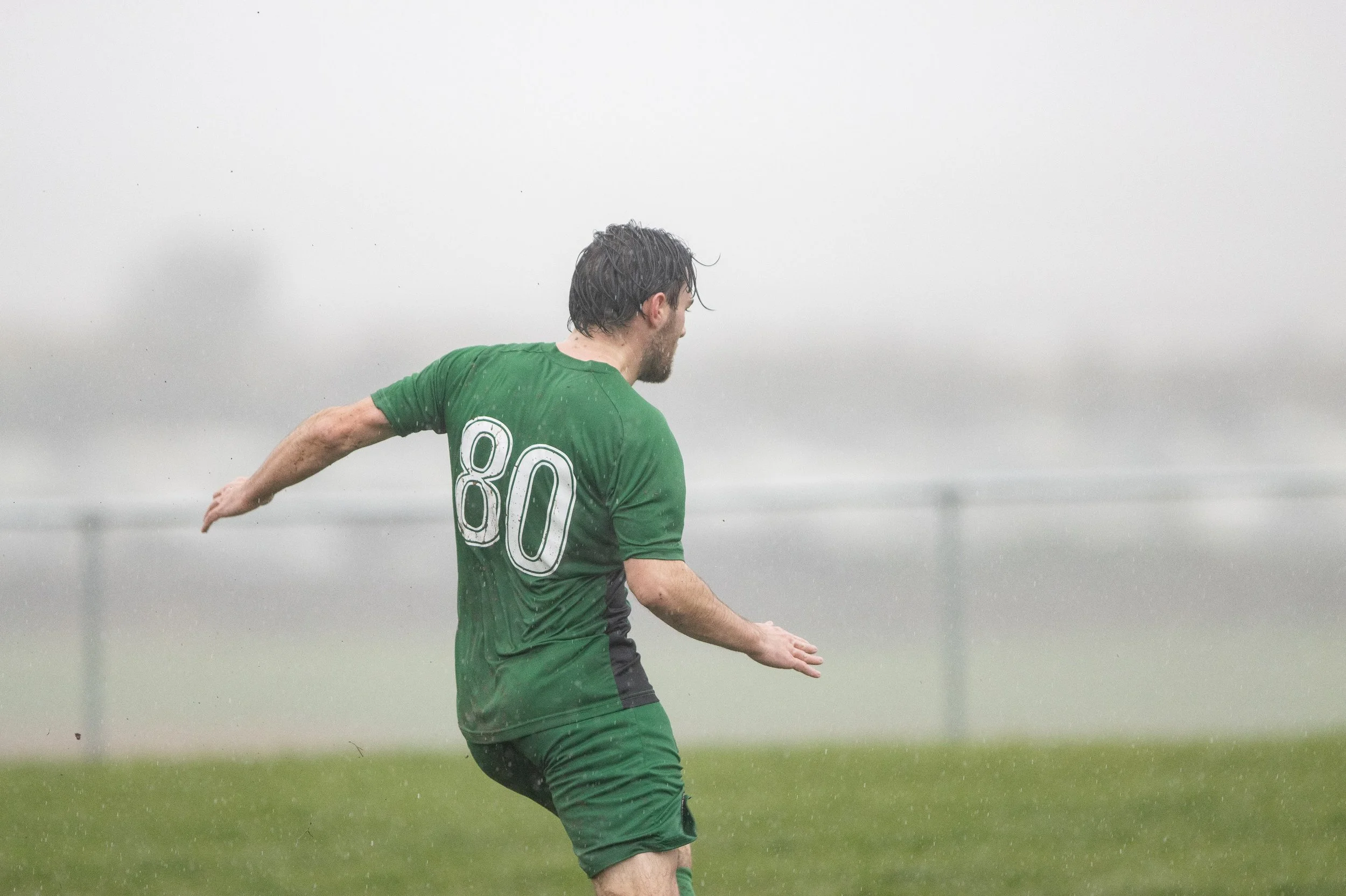A man wearing a green sports jersey with the number 80, playing soccer in the rain on a foggy field.